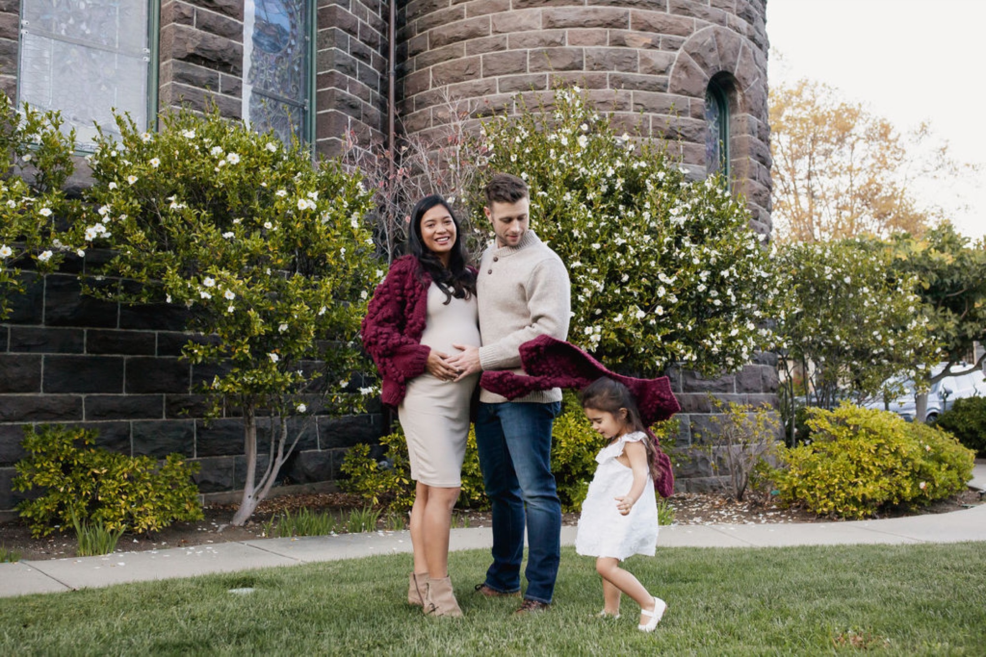 Pregnant mother in burgundy cardigan stands with husband and toddler daughter against ivy-covered brick tower on manicured lawn