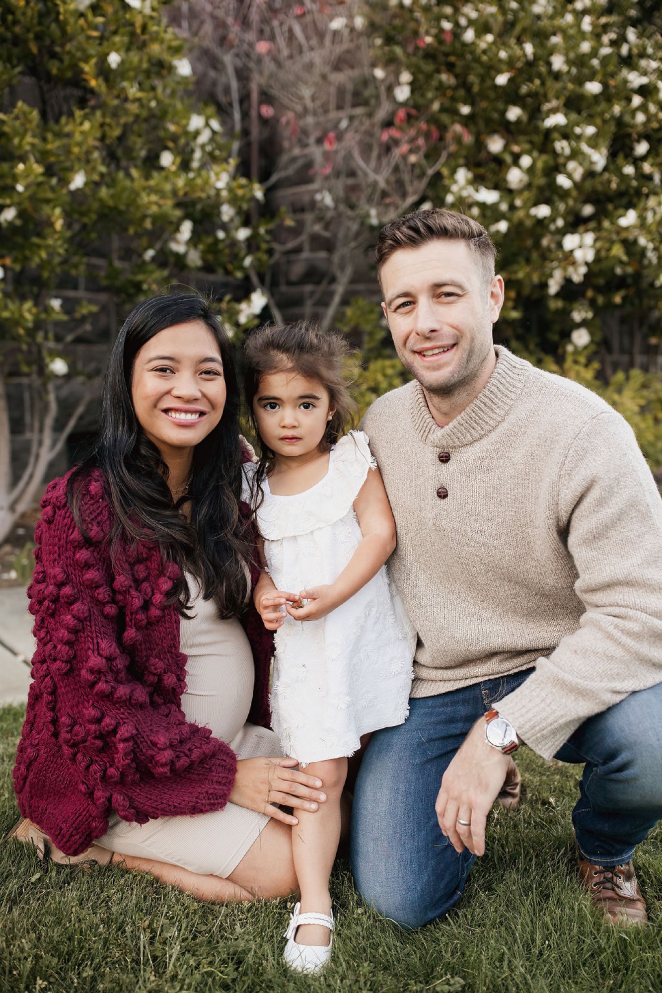 A smiling family of three kneels together on grass beneath blooming trees, the mother in burgundy and father in cream cardigan flanking their young daughter in white — San Anselmo Family Photographer