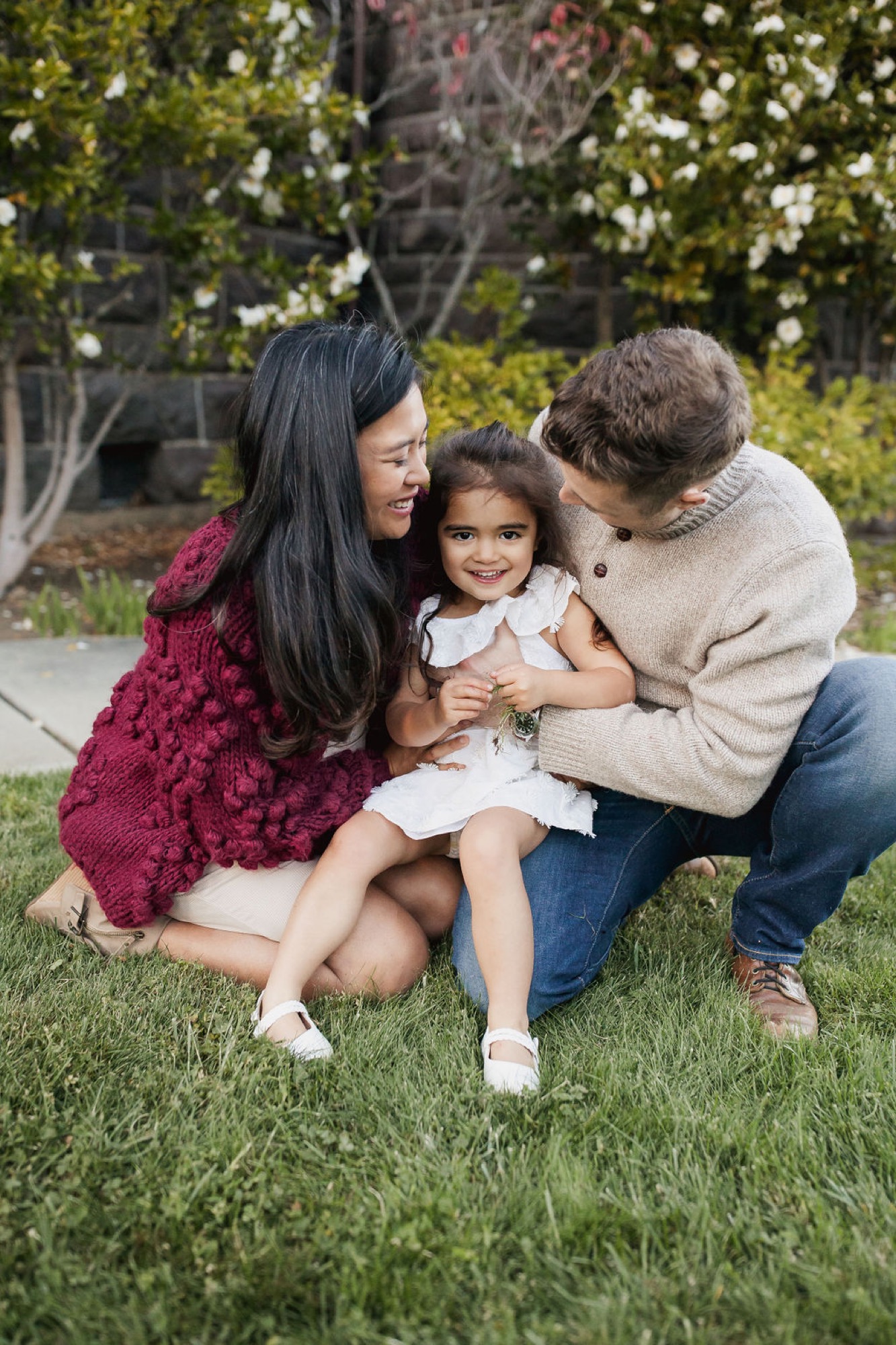 A young family crouches together on grass, with both parents embracing their small daughter who gazes downward in a moment of tender connection