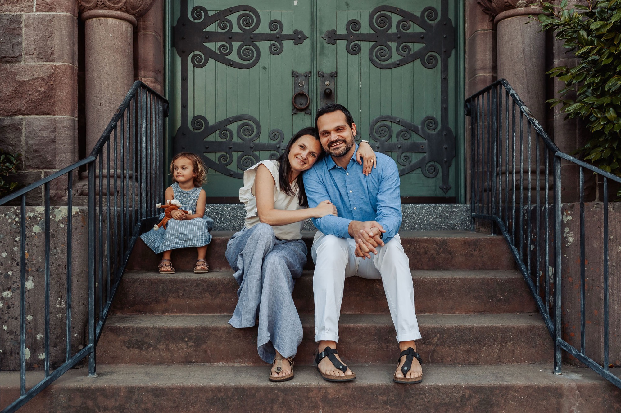 A father and mother sit closely on brick steps with their young daughter, smiling warmly at the camera beneath an ornate green door