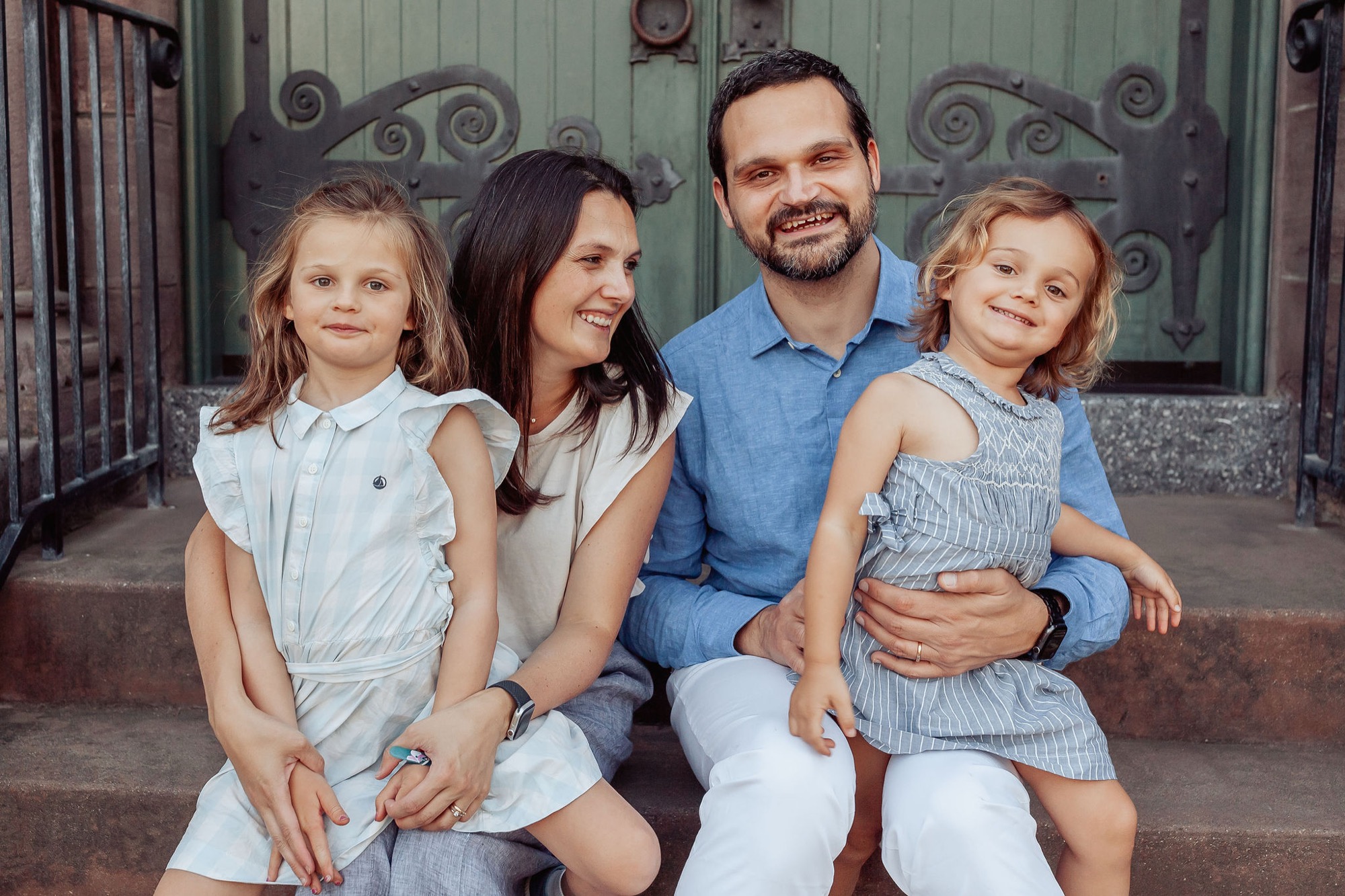 A smiling family of four sits together on a brick step against an ornate green gate, with two young girls in light blue outfits flanking their parents