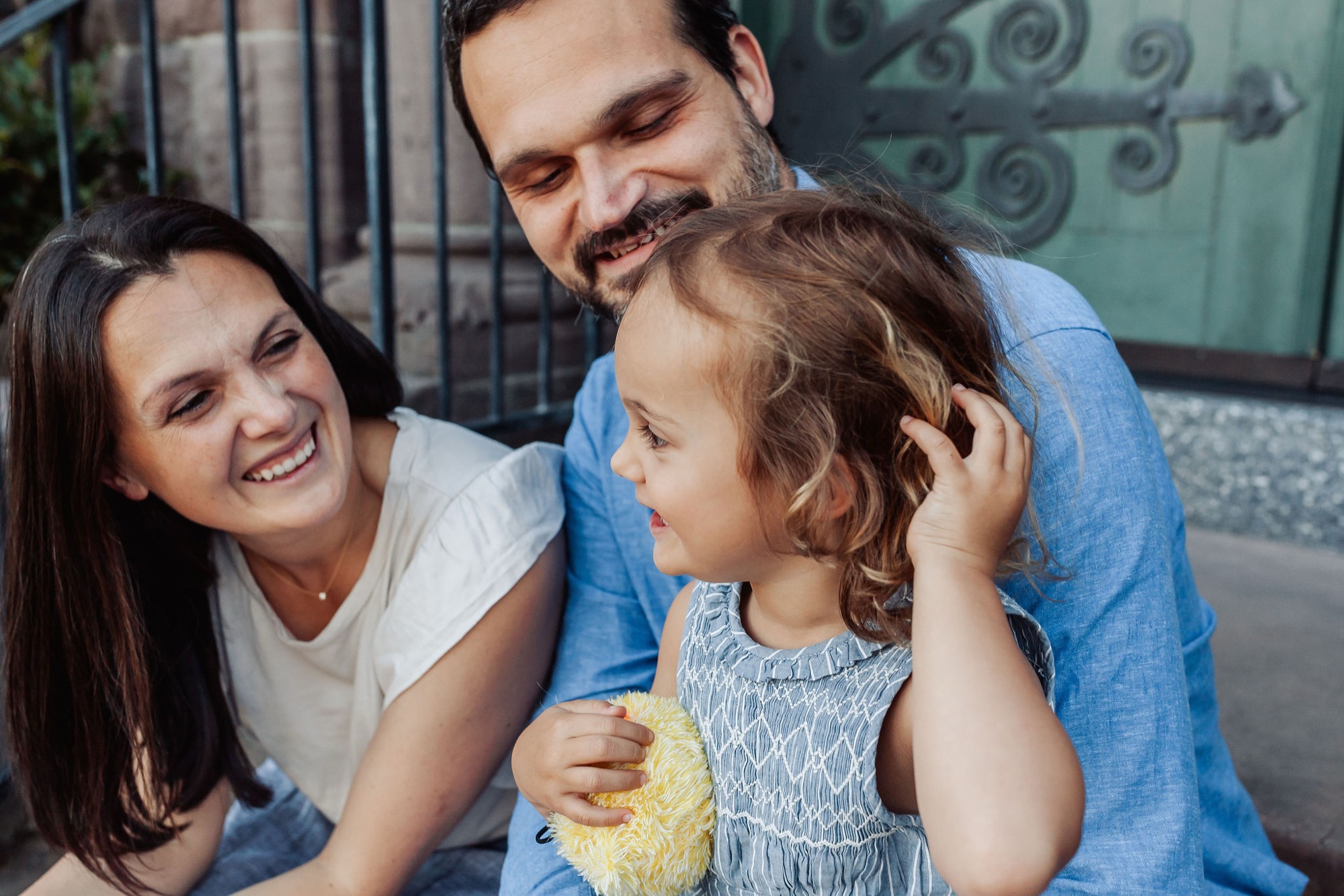 Parents lean in with genuine smiles as their young daughter touches her father's face in a moment of playful affection against an ornate green door