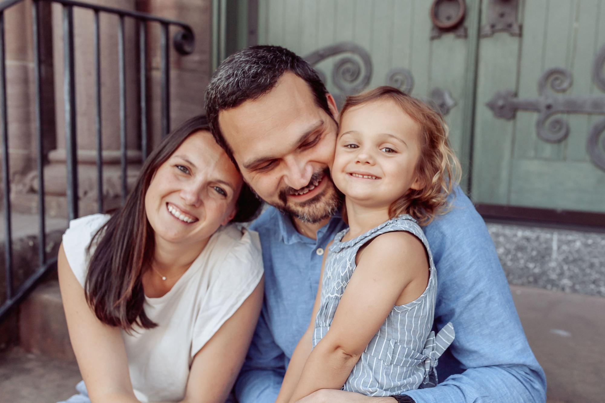 A smiling father in a blue shirt hugs his wife and young daughter against a weathered green door, all three beaming with genuine joy