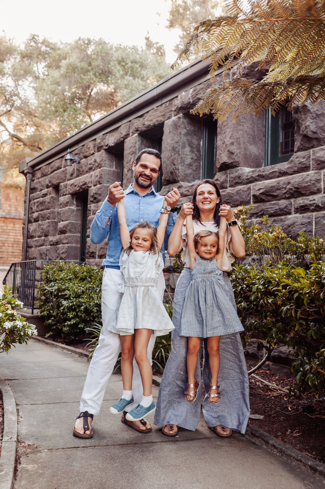 A family of four stands cheerfully on a concrete path in front of a rustic stone building, with warm afternoon light filtering through surrounding foliage