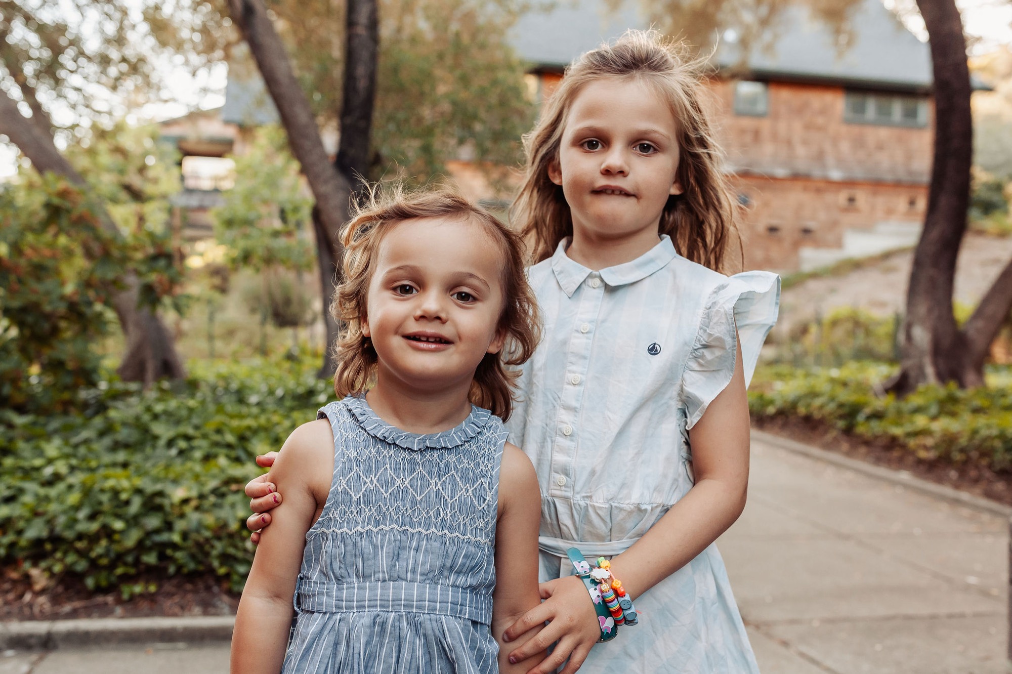 Two young girls hold hands while standing on a tree-lined residential path, both smiling directly at the camera with natural afternoon light filtering through the canopy