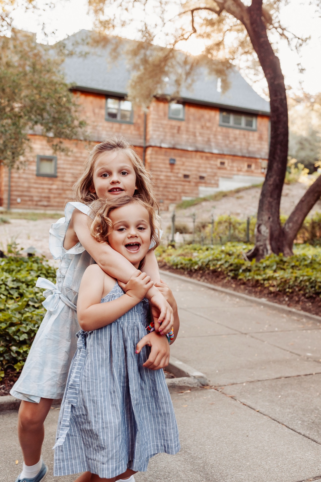 Two young girls in light blue dresses embrace on a sidewalk, their genuine smiles radiating joy beneath a sprawling tree