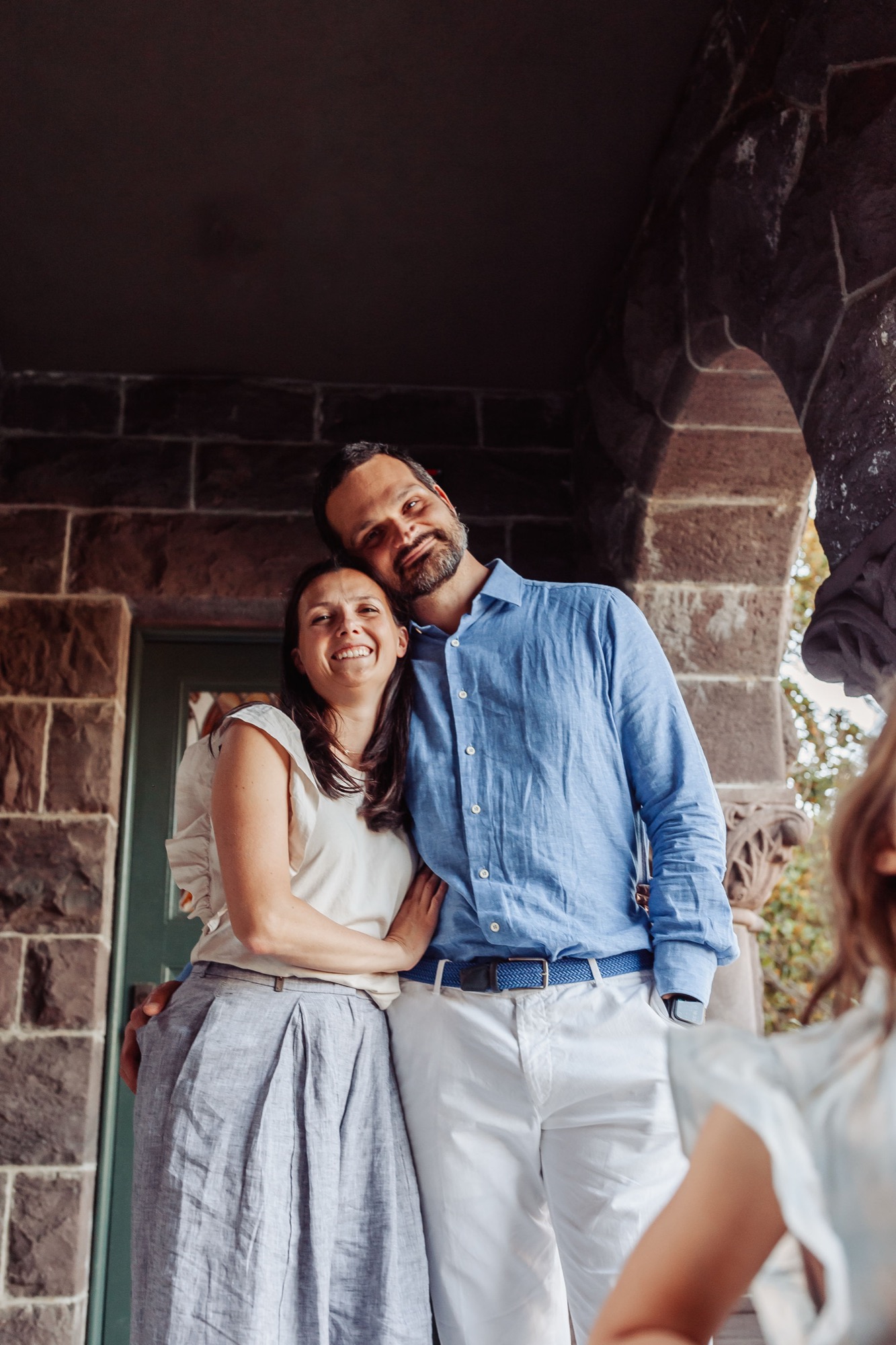 A couple stands close together in the doorway of a stone building, warmly smiling at the camera as natural light frames their relaxed embrace
