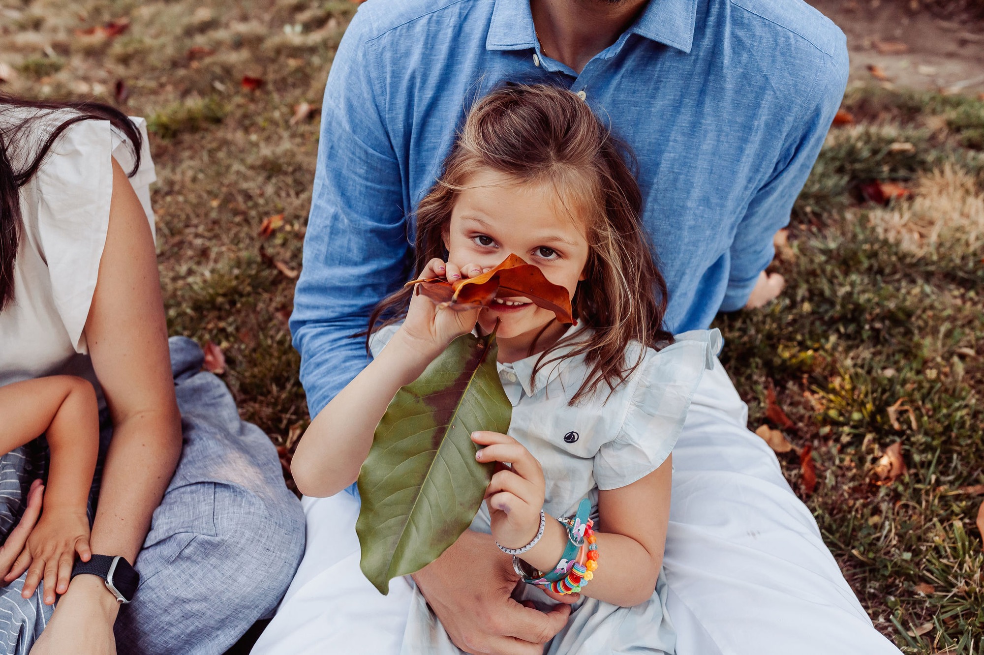 A young girl holds autumn leaves to her face while sitting with her father on grass, her expression playful and curious