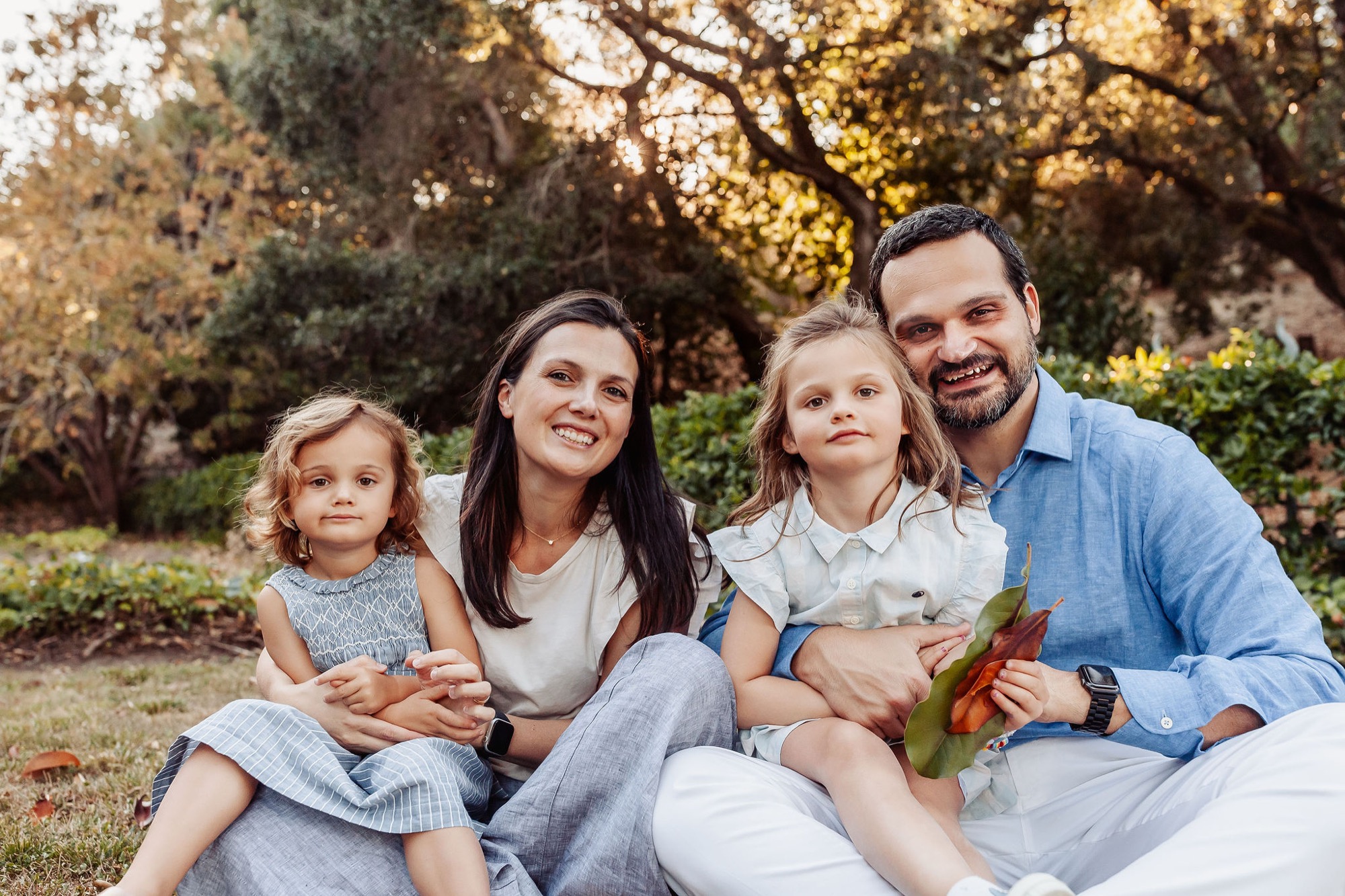 Family of four sits together on grass beneath golden autumn trees, smiling warmly at the camera in afternoon light