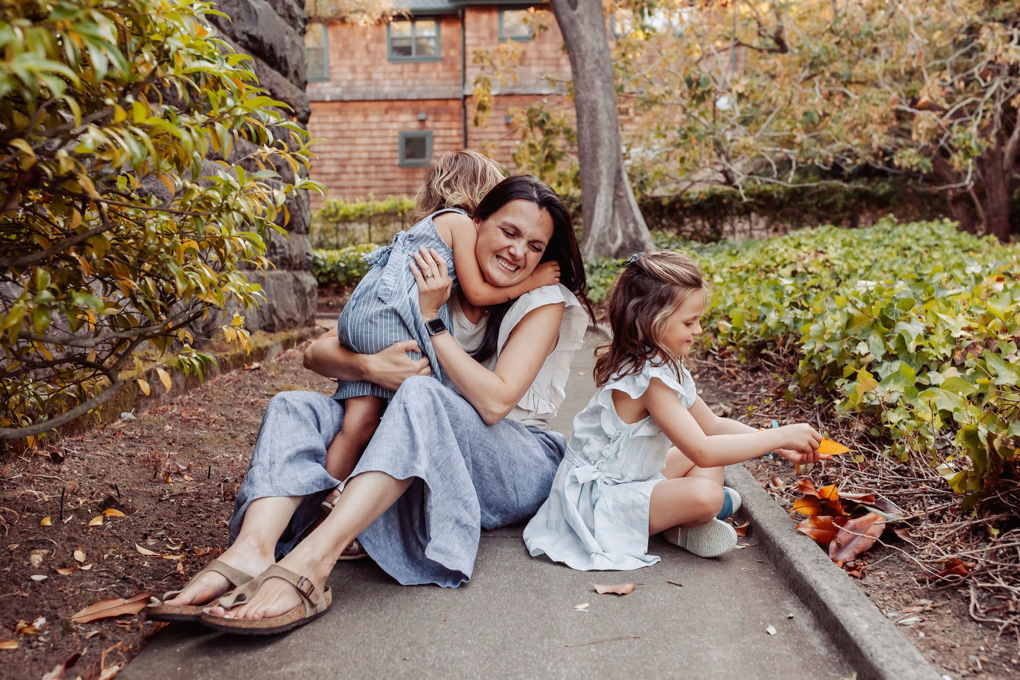 Mother holds her young son while sitting beside her daughter on a shaded garden path, all three smiling at the camera