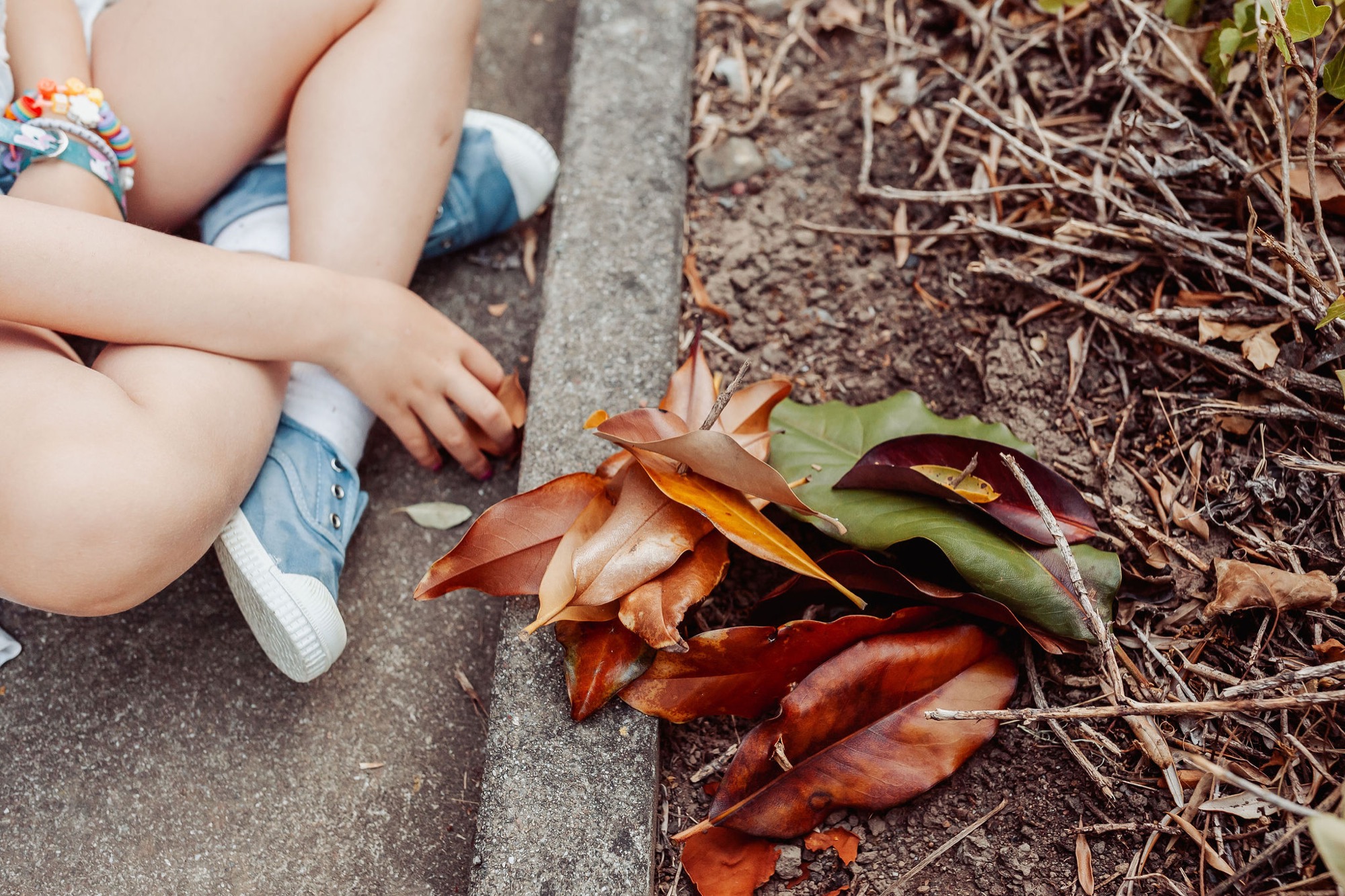 A child's bare legs dangle as small hands reach down to examine fallen autumn leaves scattered on concrete