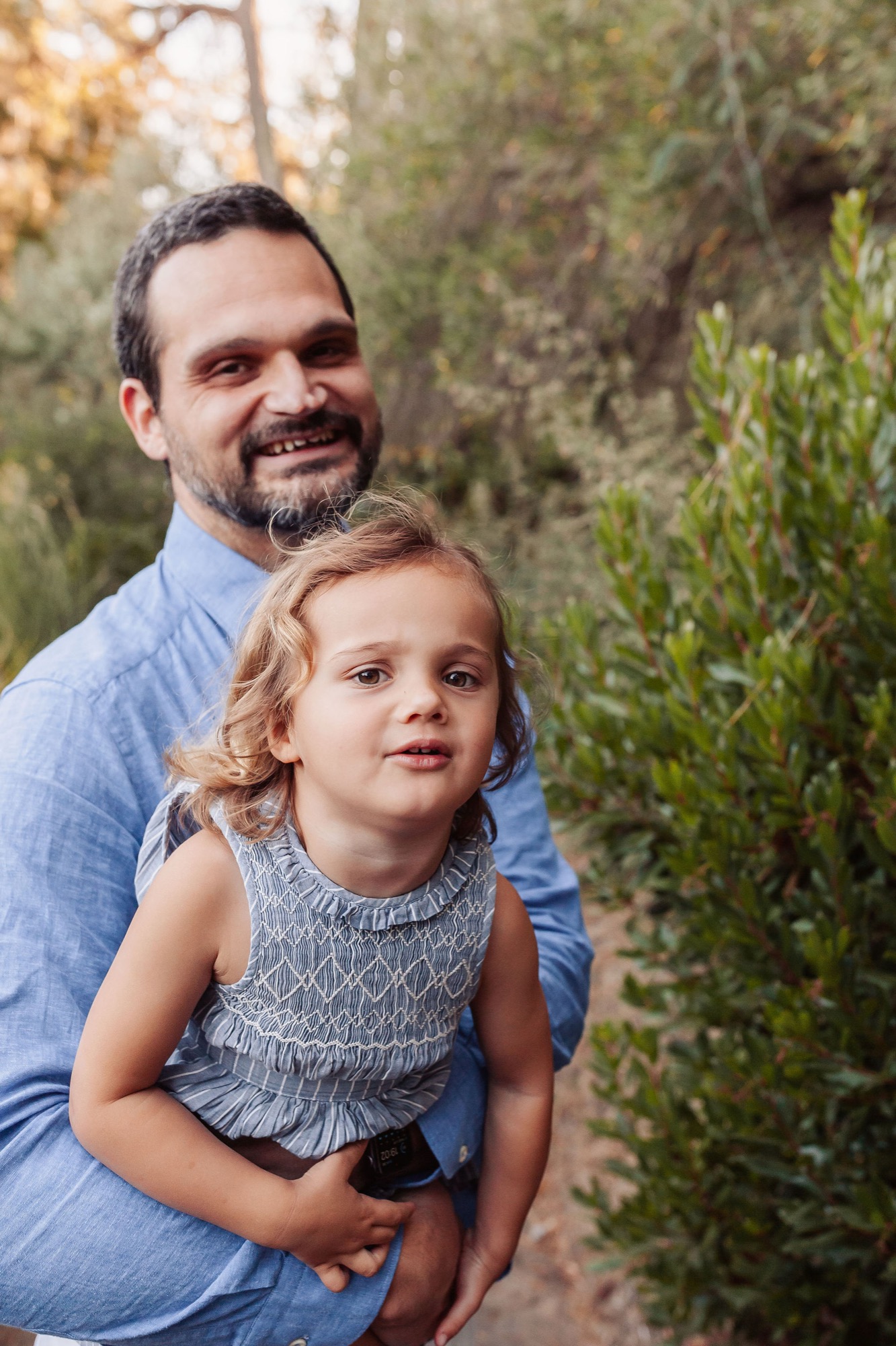 A bearded father in a blue shirt holds his young daughter with blonde curls against evergreen shrubs, both gazing thoughtfully to the side