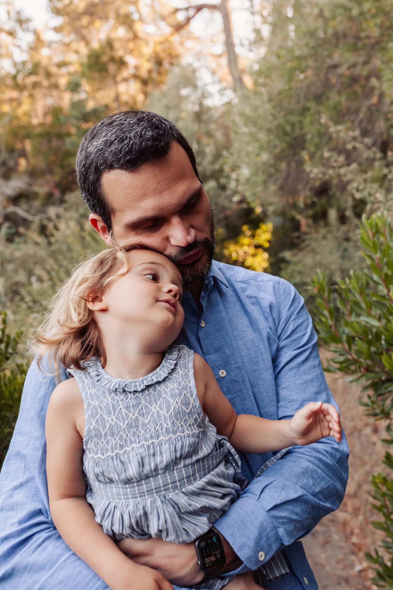 A bearded father holds his young daughter while gazing upward together among lush green foliage in dappled natural light