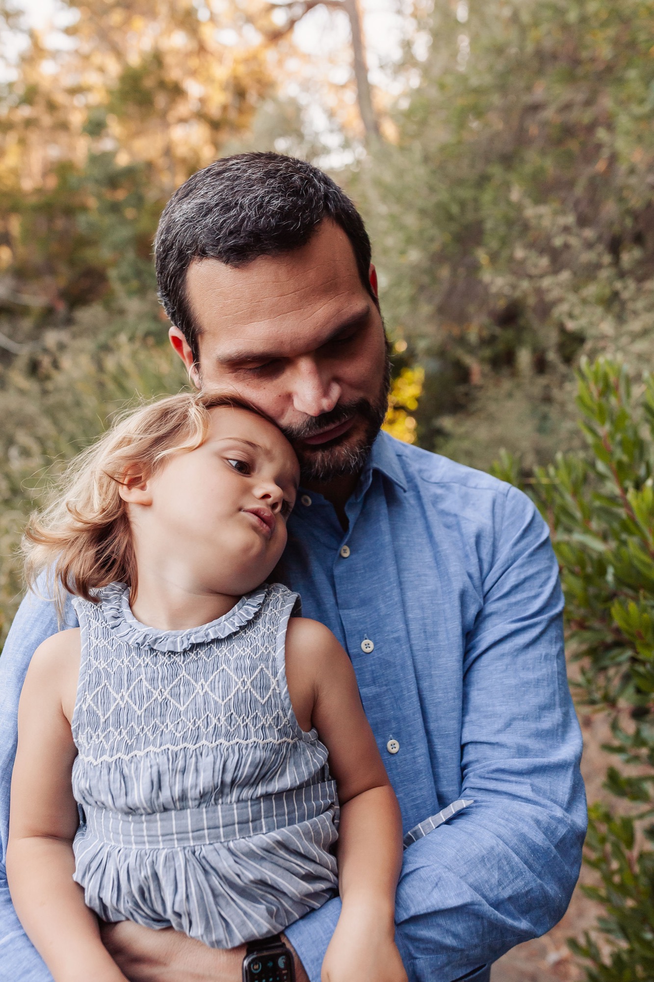 A bearded father gazes down tenderly at his young daughter in a gray smocked dress, both framed by soft natural foliage and dappled autumn light