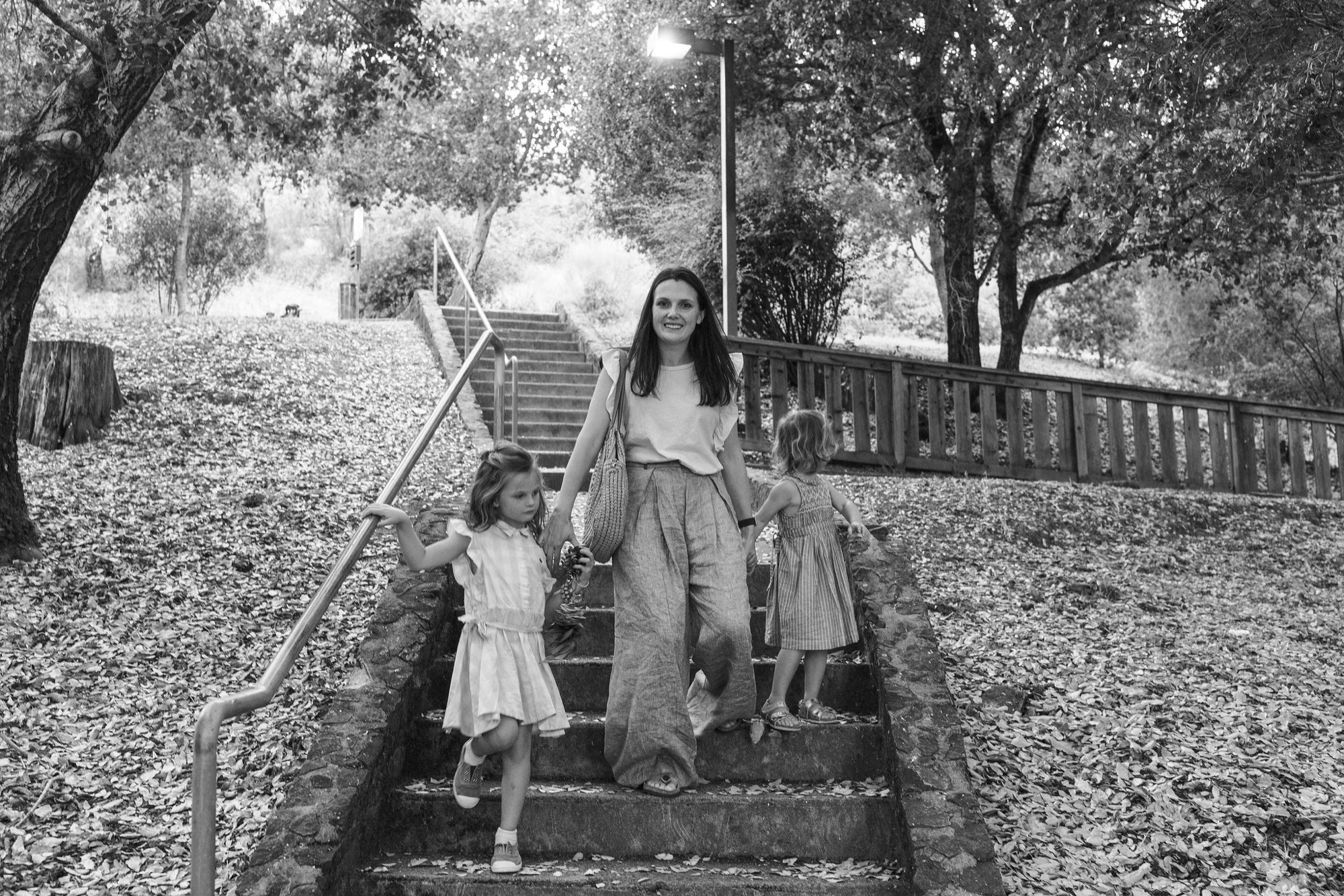 Woman and two young girls descend wooden stairs through dappled forest light, the children's white dresses bright against dark tree trunks