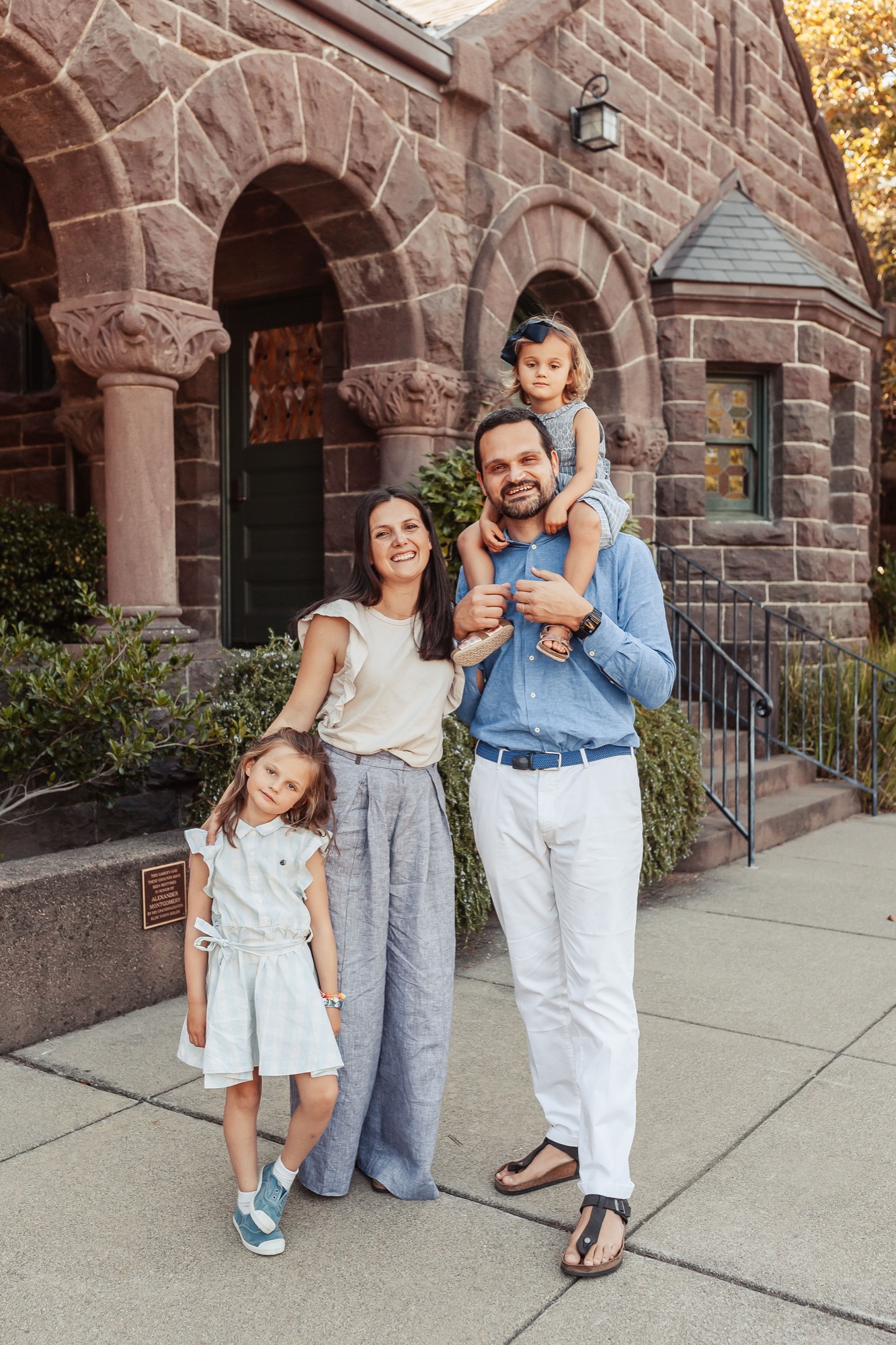 Family of four poses on a sunny sidewalk before a historic red-stone building with arched doorways and climbing ivy