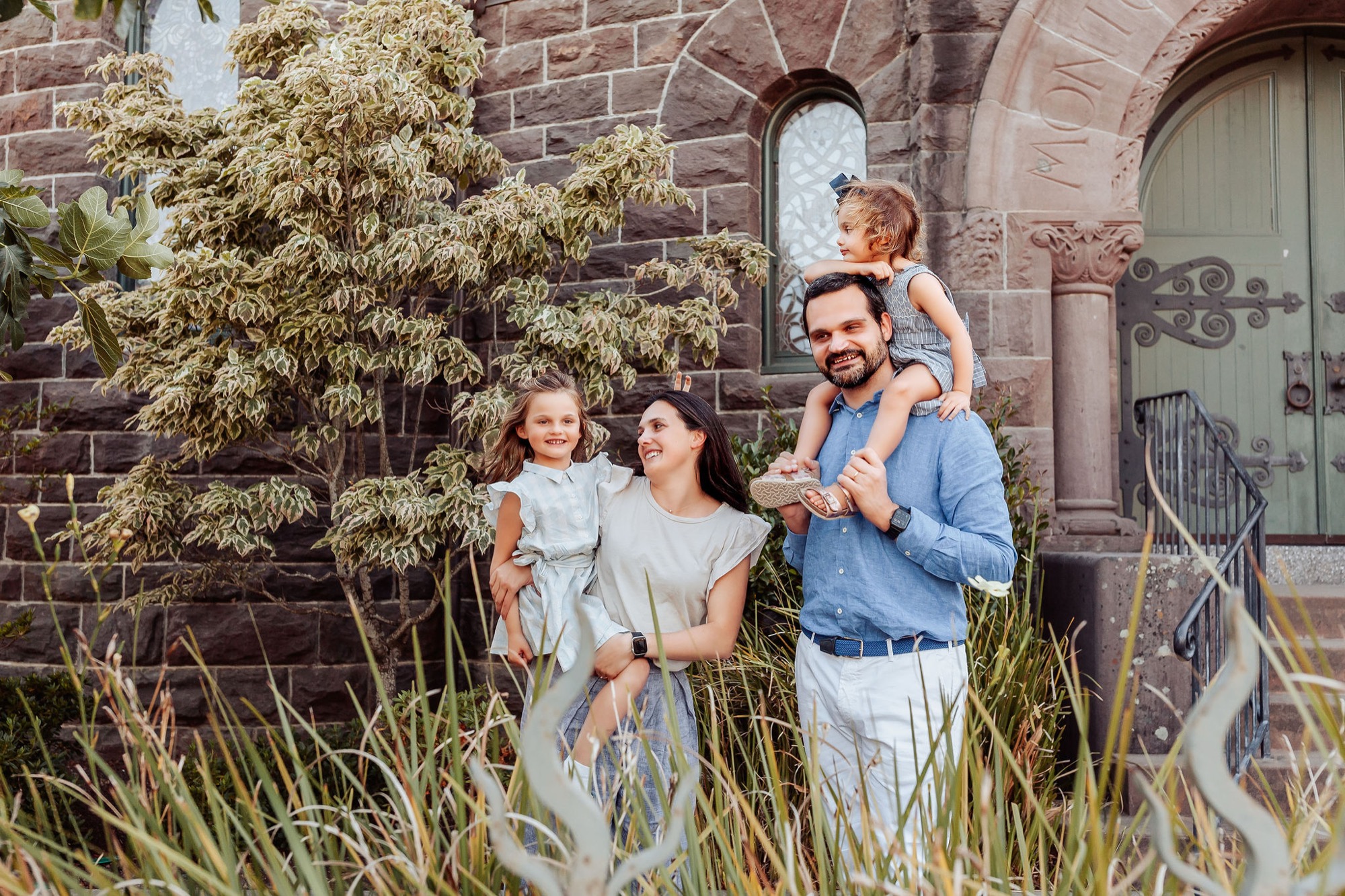 Family of four stands together against a brick church building, father holding son on shoulders while mother and daughter smile warmly in soft afternoon light