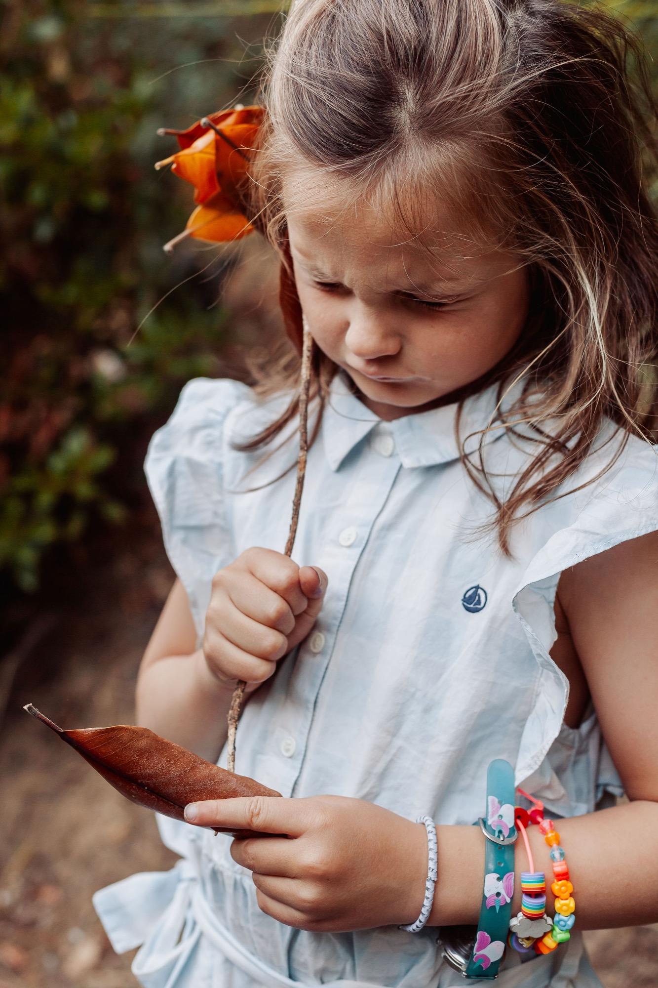 Young girl examines a fallen autumn leaf with focused concentration, wearing a light blue shirt and colorful beaded bracelets