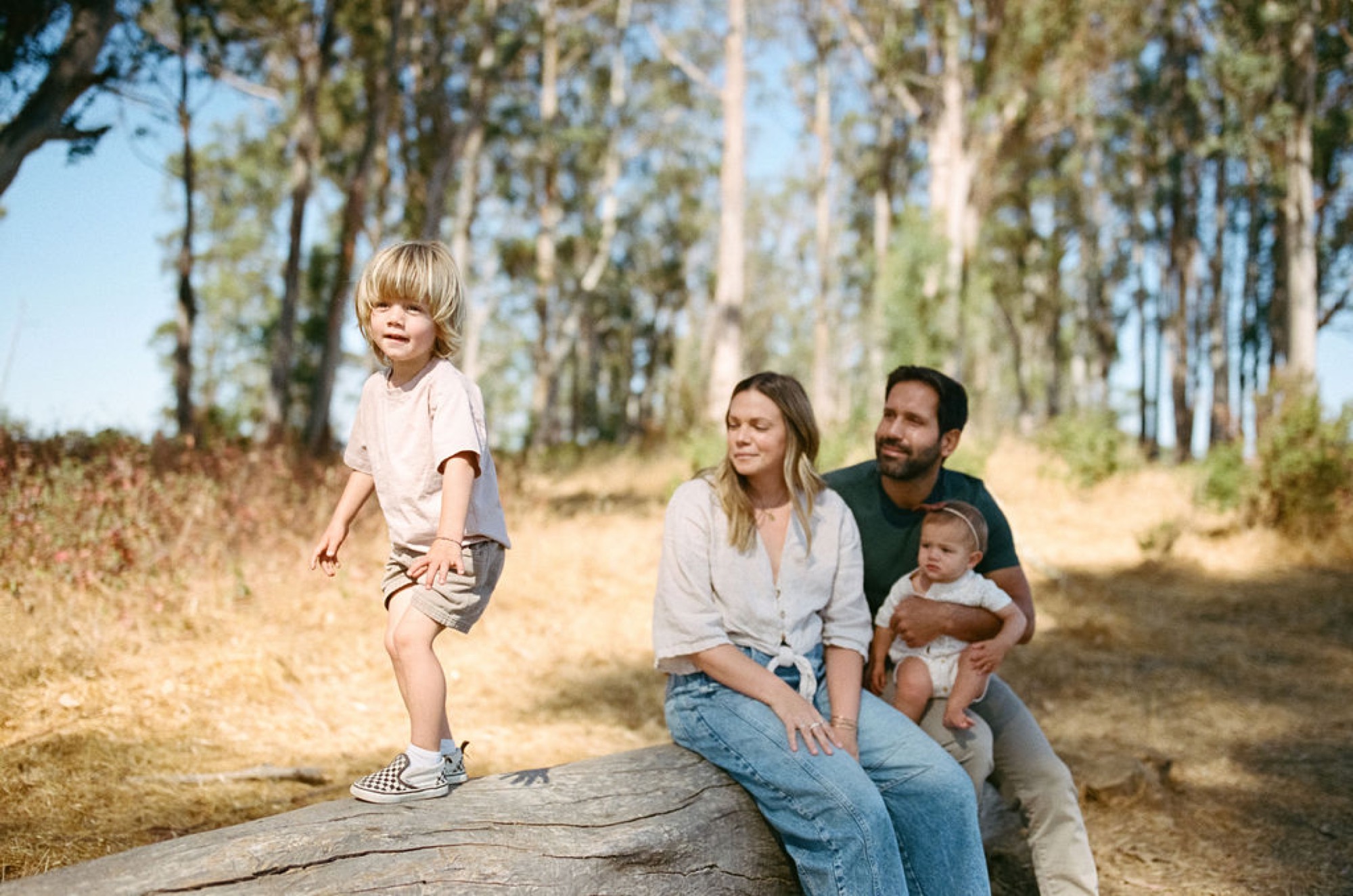 Young boy in checkered sneakers balances on a fallen log while parents watch, tall eucalyptus trees framing the dusty trail behind them