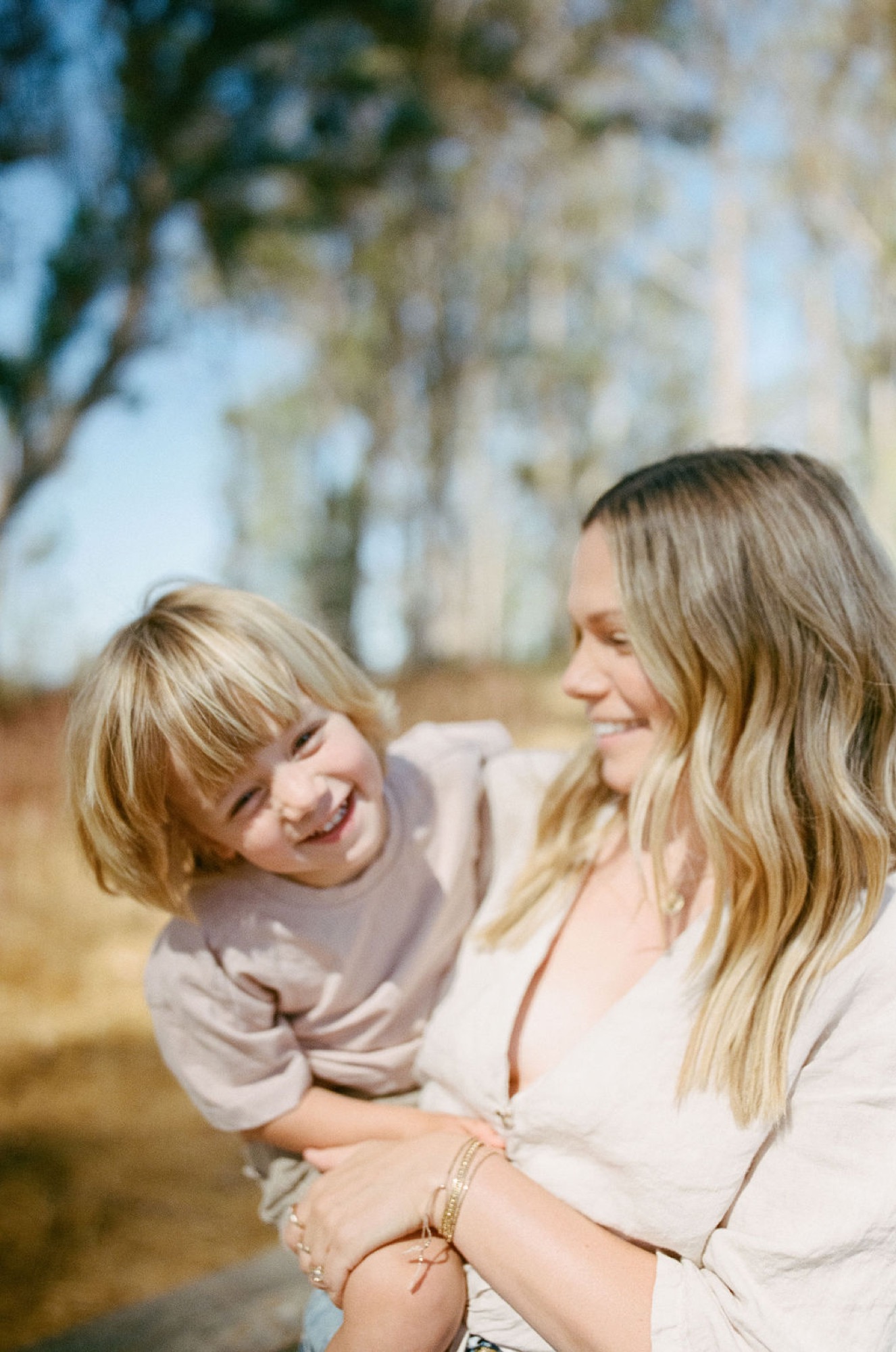 A woman holds her laughing toddler in warm afternoon light beneath blooming white flowers, both radiating joy