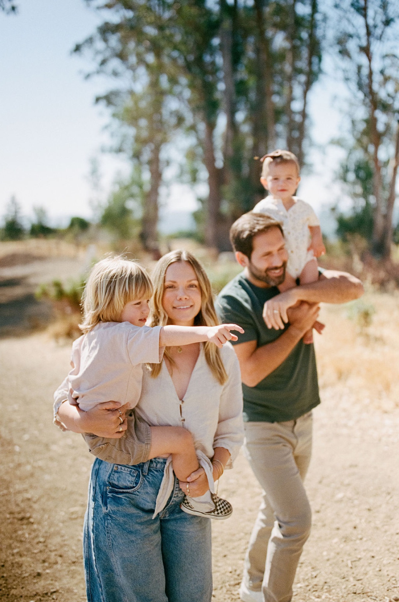 A family of four walks together on a dusty trail lined with towering eucalyptus trees, with warm golden sunlight illuminating their relaxed smiles and casual embrace