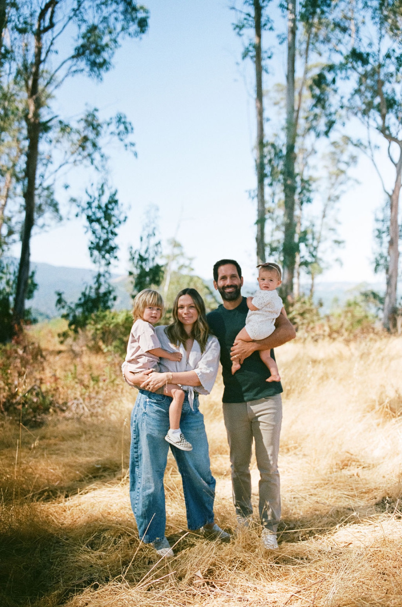 Family of four posing together on a golden grass hillside among tall trees at the open space preserve in downtown San Rafael