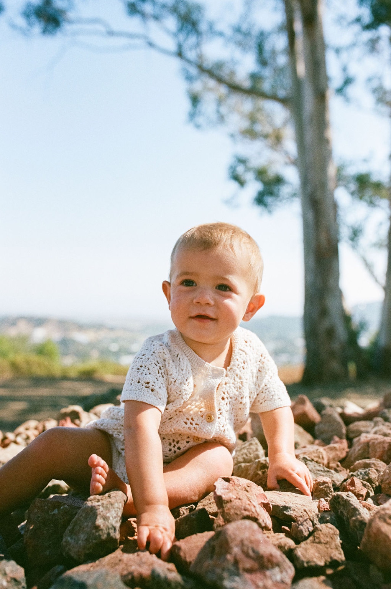 A blonde toddler in a cream knit shirt sits among rust-colored rocks, gazing thoughtfully toward the camera with a cypress tree and valley vista behind