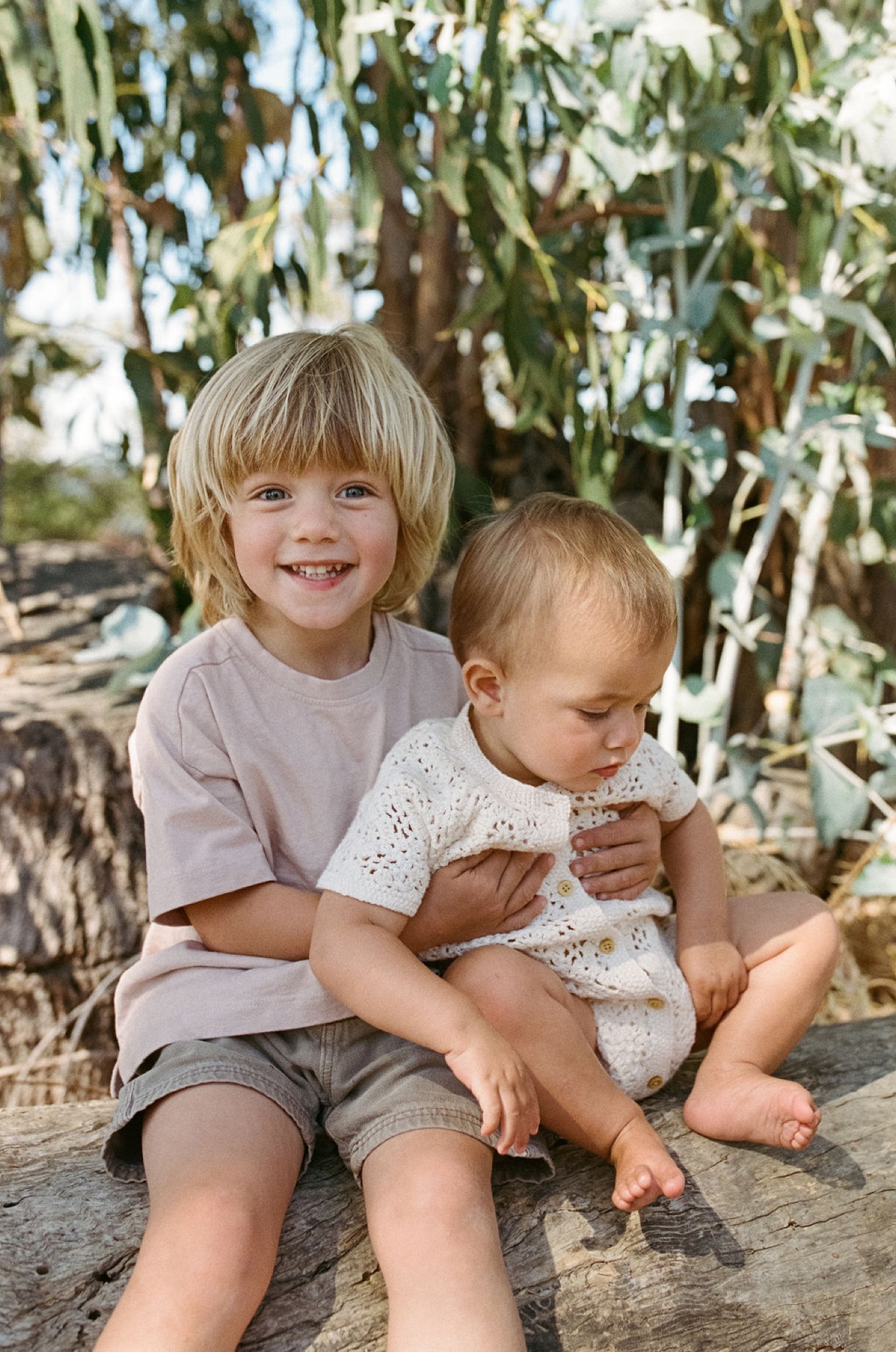 Older sibling beams at camera while cradling barefoot toddler on weathered stone beneath drooping willow branches