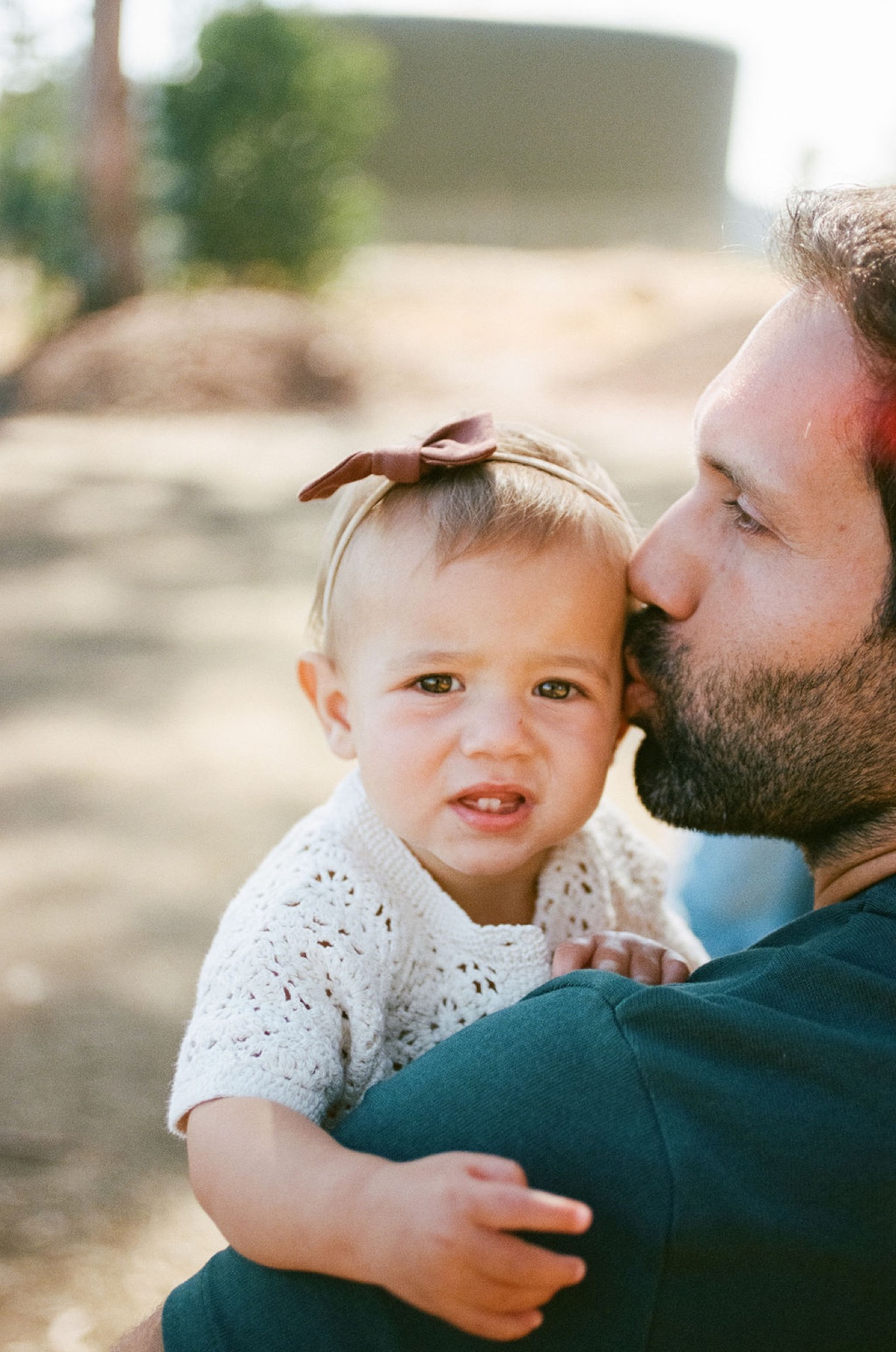 A bearded father tenderly kisses his toddler's head while holding her close, her bright eyes gazing toward the camera with soft afternoon light illuminating their faces