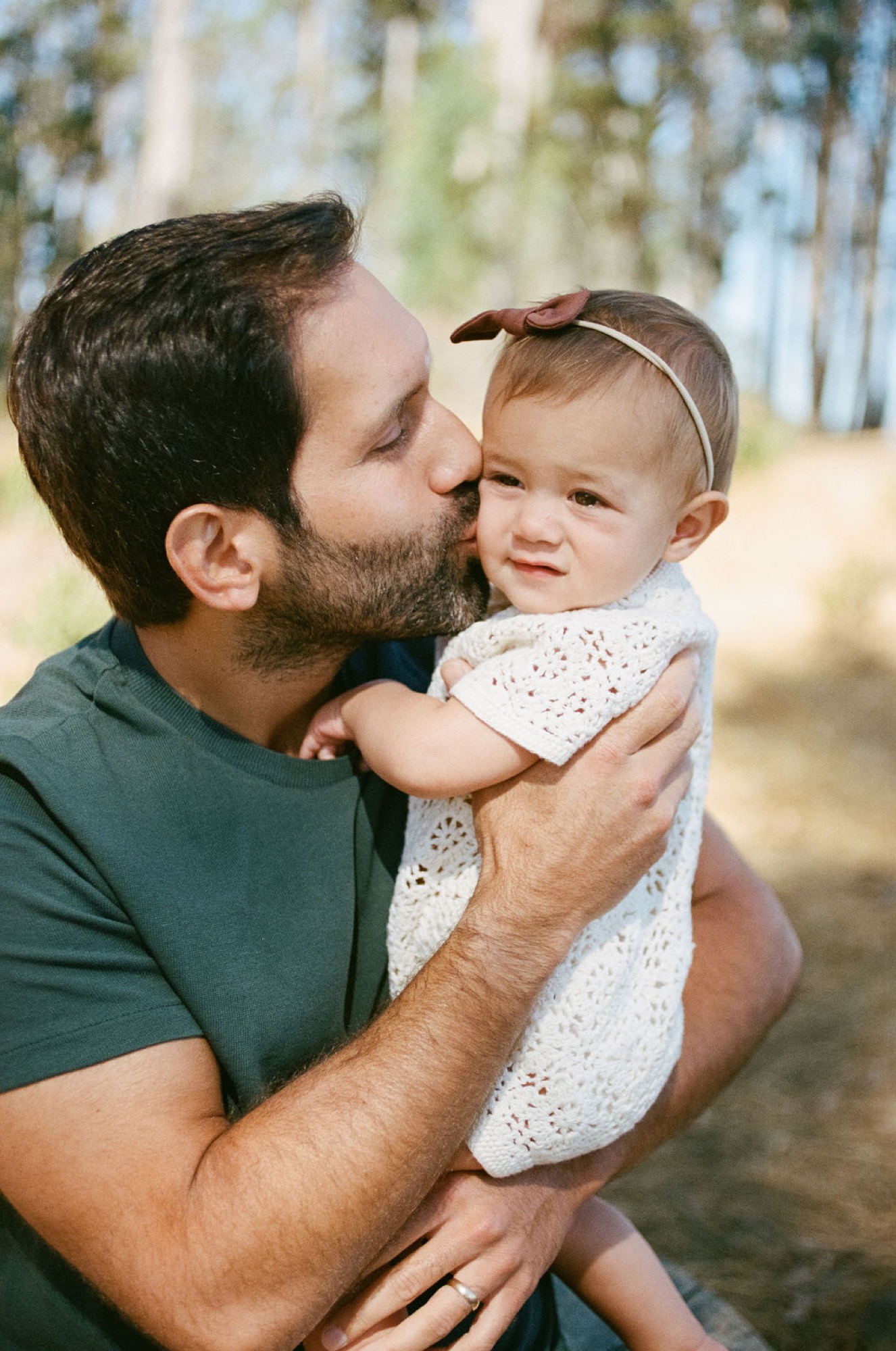 A bearded father kisses his toddler's cheek while holding her against his chest, dappled sunlight filtering through tall trees behind them