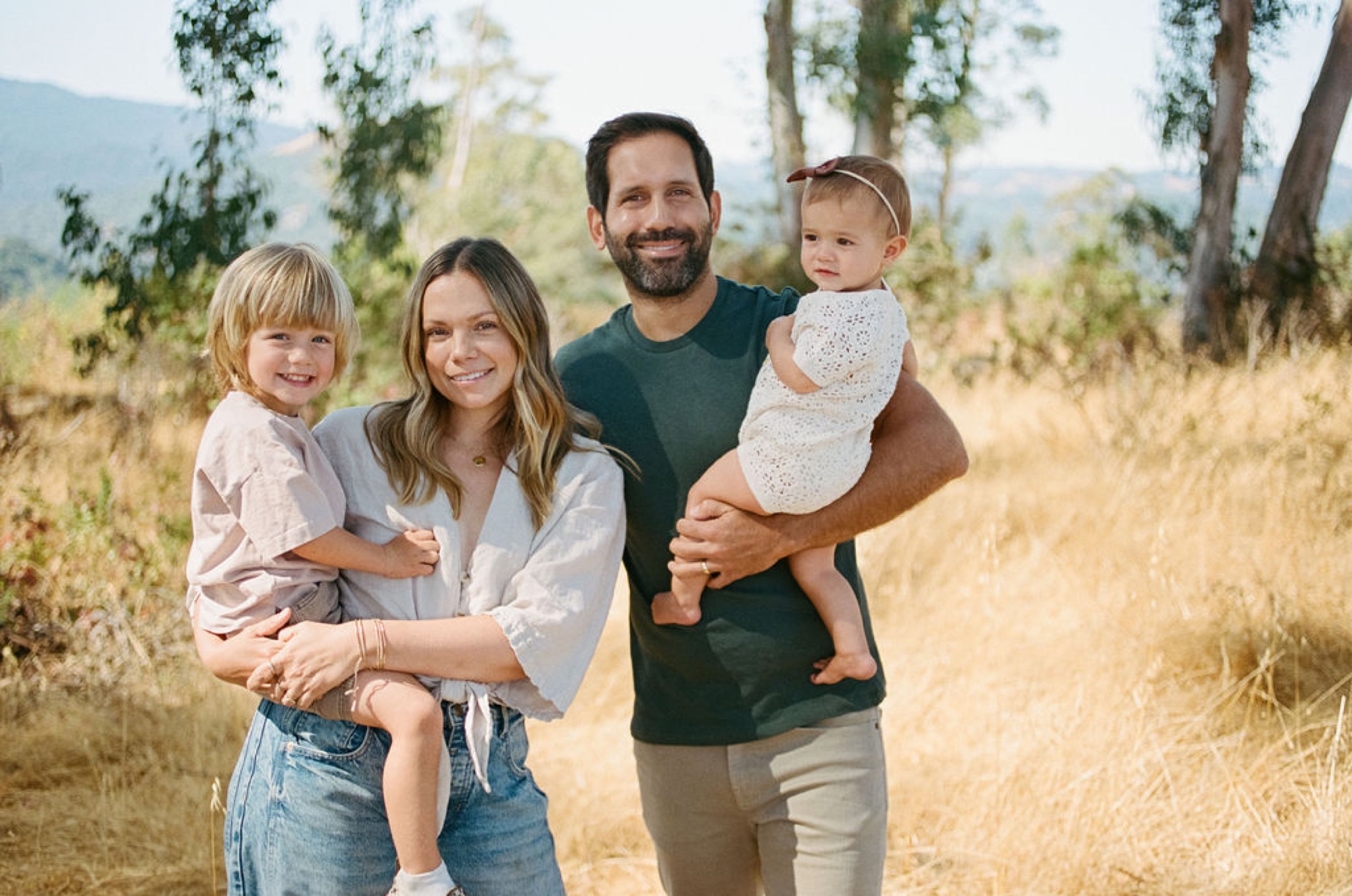 A smiling family of four stands on a sun-dappled trail, with the father holding their youngest child while the mother embraces their older son, mountains visible in the soft background