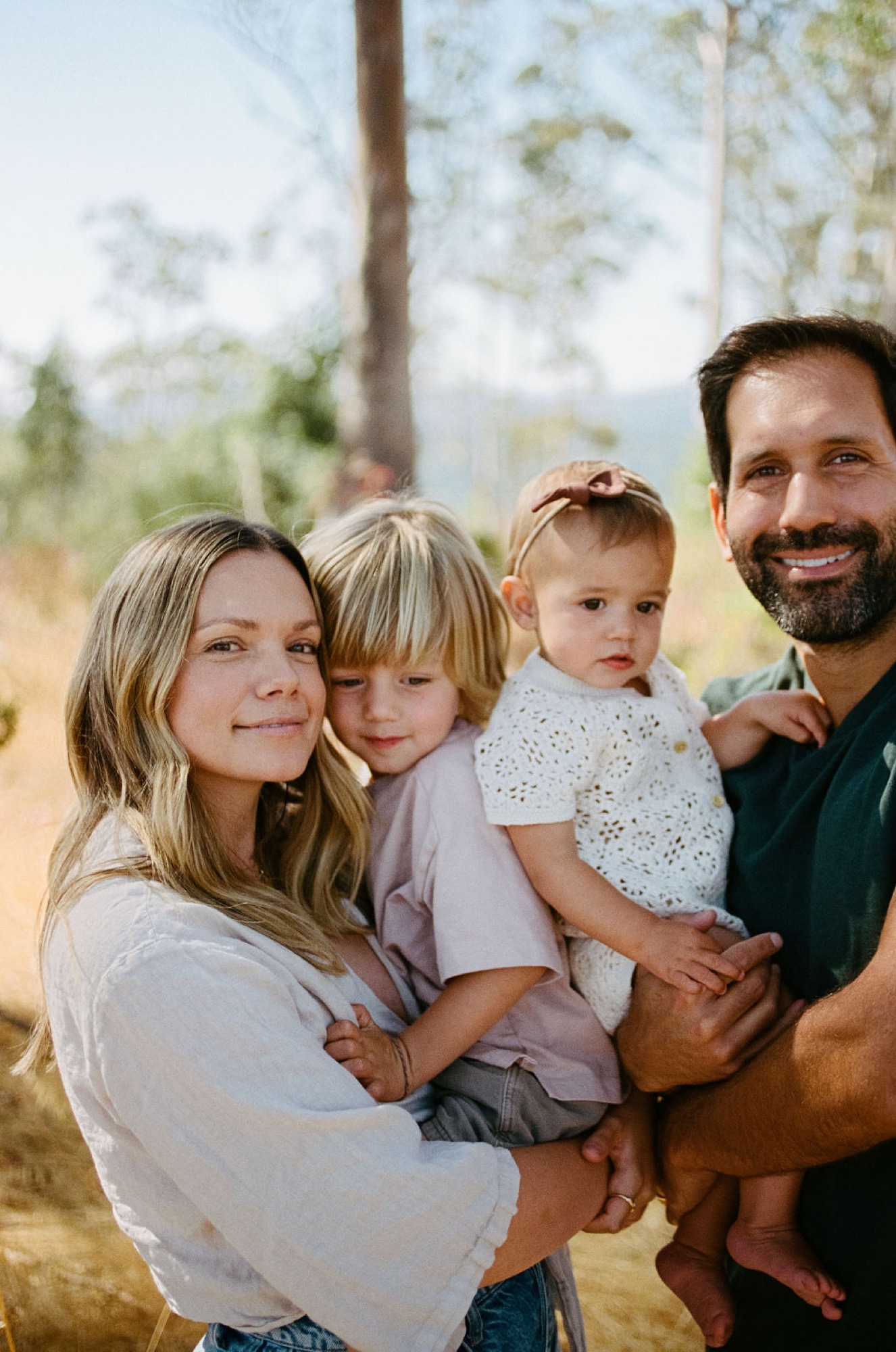 A family of four smiles warmly at the camera beneath tall pines, golden afternoon light filtering through the trees behind them