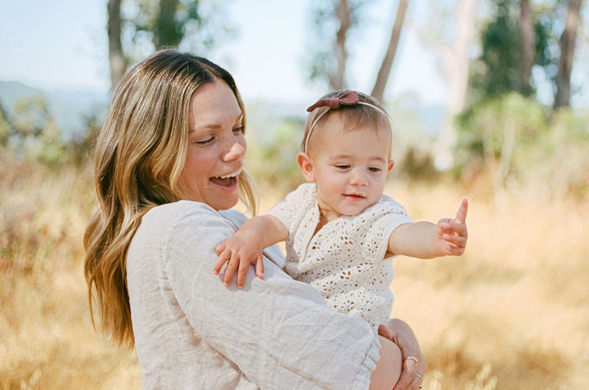 A woman in a light gray shirt holds her toddler daughter in an open field, both smiling as the child reaches outward with extended arms