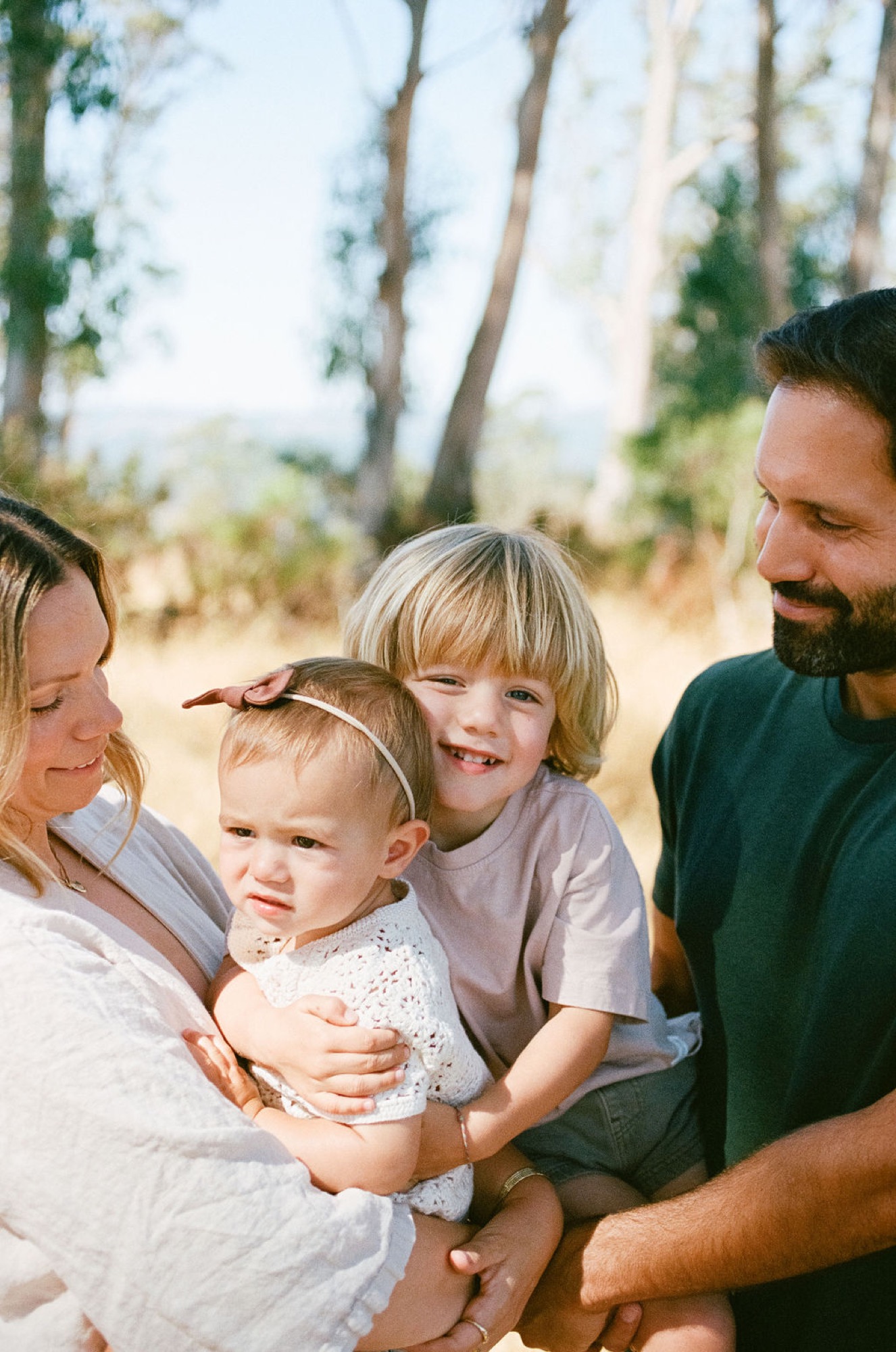 A family of four gathers in dappled sunlight beneath tall trees, the toddler nestled between parents while an older sibling beams with genuine joy
