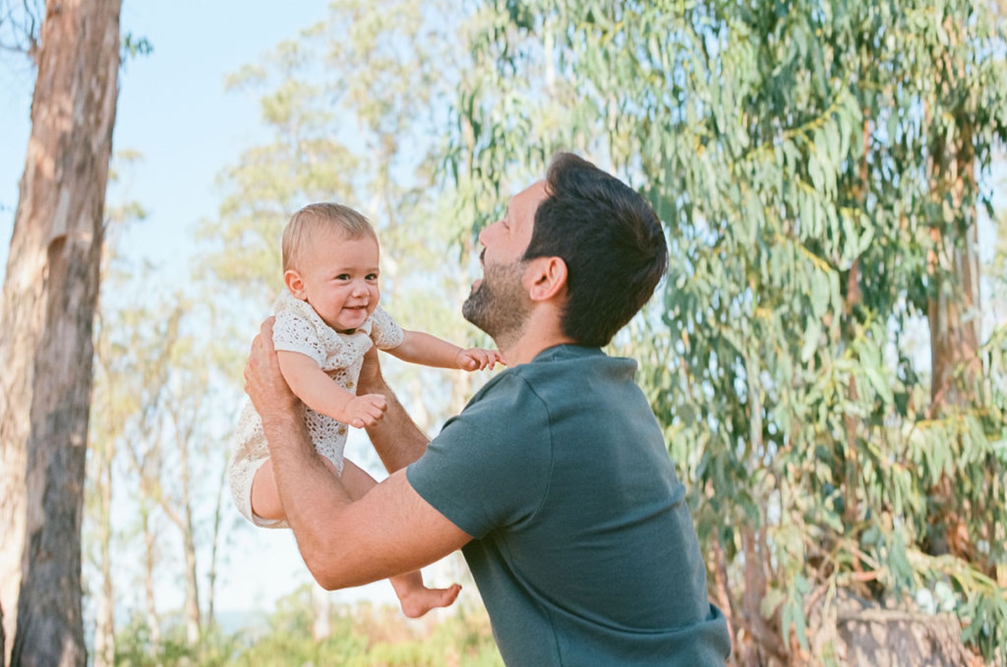 A bearded father lifts his smiling toddler against tall eucalyptus trees, dappled sunlight filtering through the canopy above