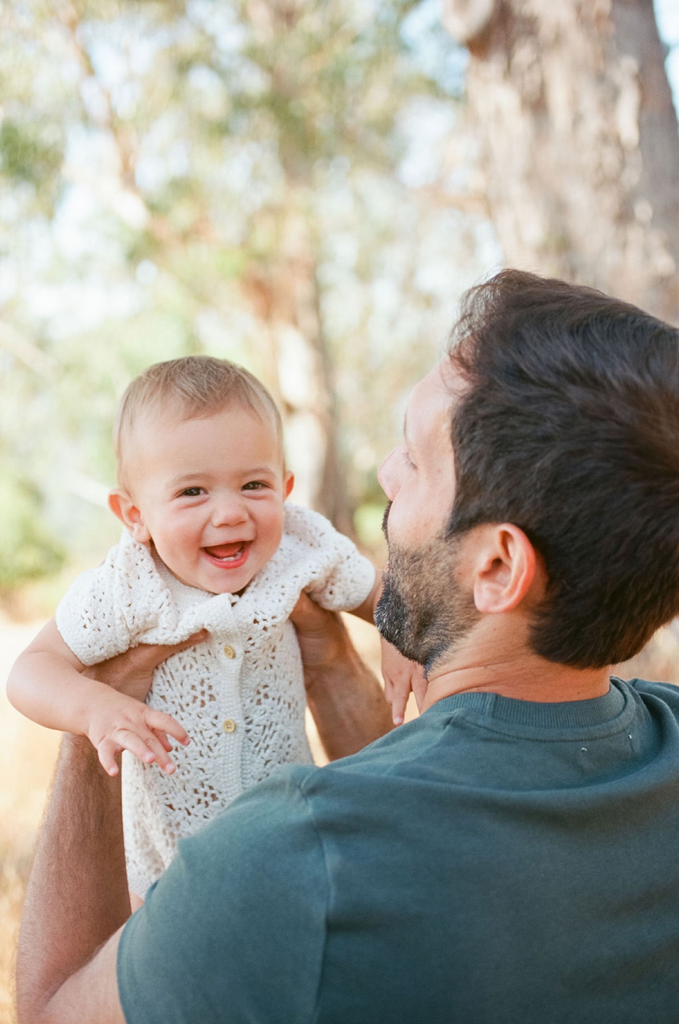 A bearded father holds his laughing infant in dappled sunlight beneath blossoming trees, both radiating pure joy
