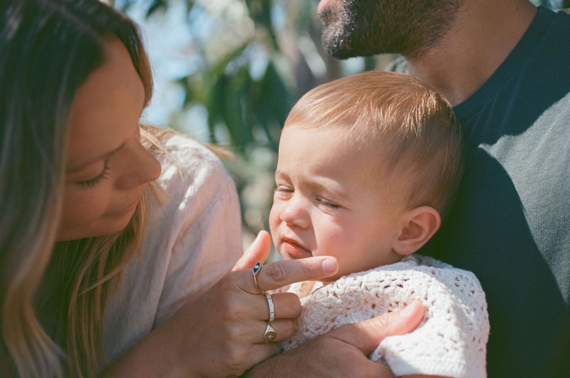 A toddler cries while cradled between two parents under dappled sunlight, a real moment from a family session at San Rafael open space, Marin County