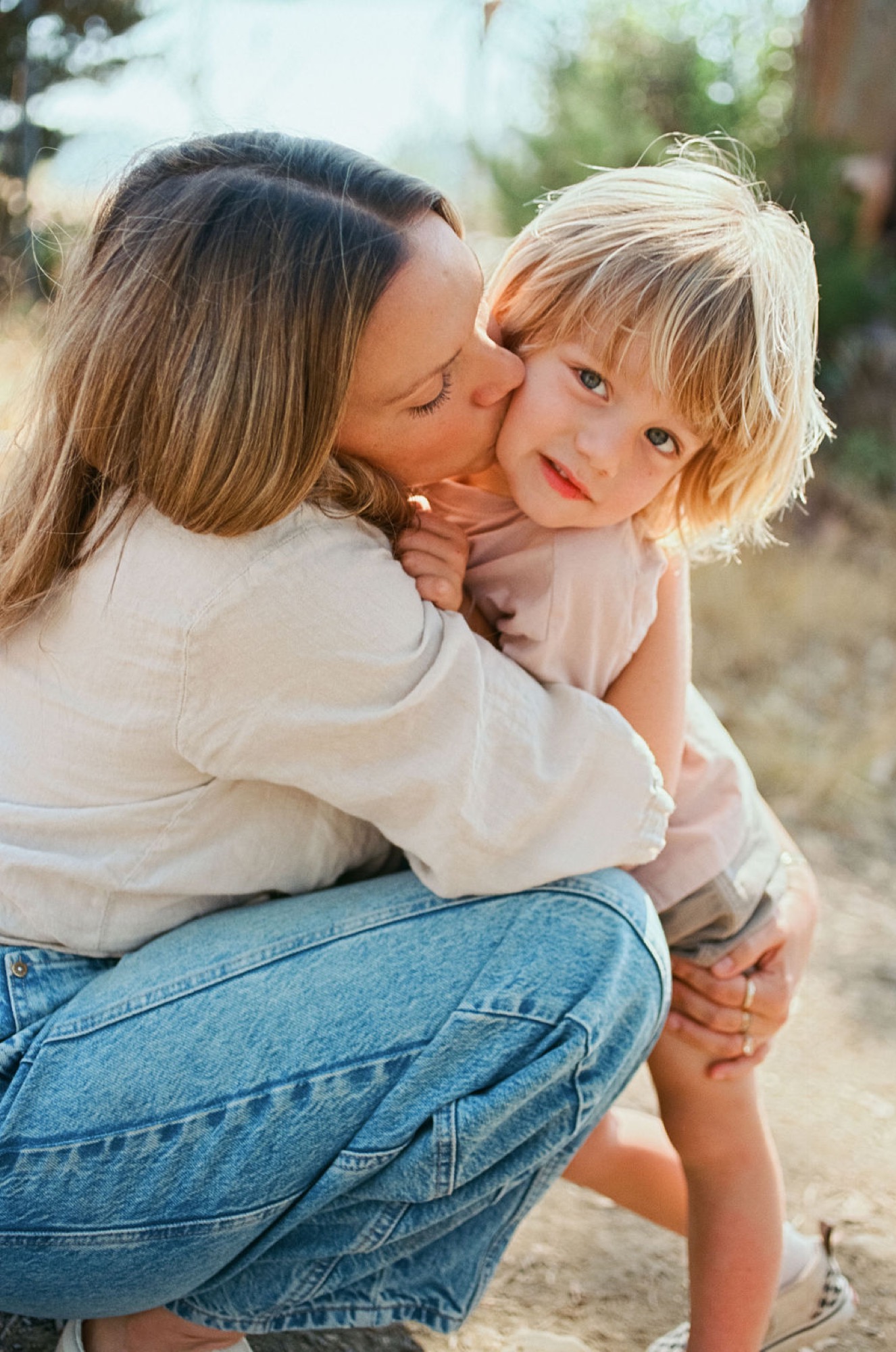 A mother kisses her young daughter's cheek while holding her in golden afternoon light, the child gazing toward the camera with a gentle expression