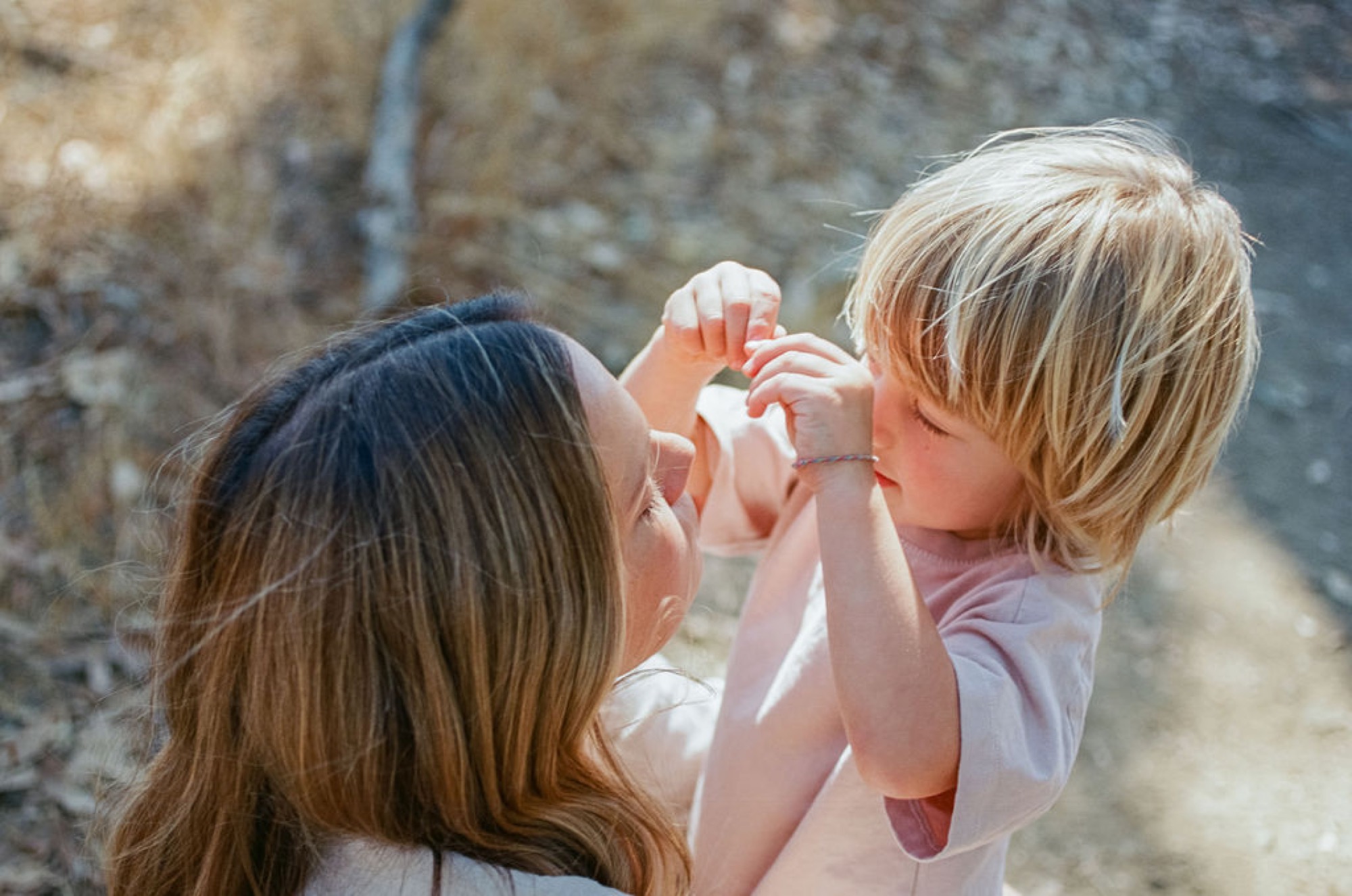 A woman lifts a blonde toddler toward her face while sitting against weathered stone, their hands meeting in joyful connection
