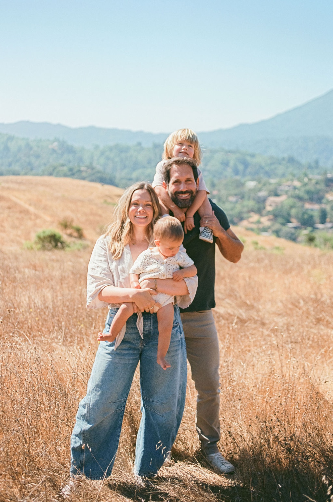 A bearded father holds his blonde toddler on shoulders while the mother smiles warmly, cradling their infant in a sun-drenched harvested field