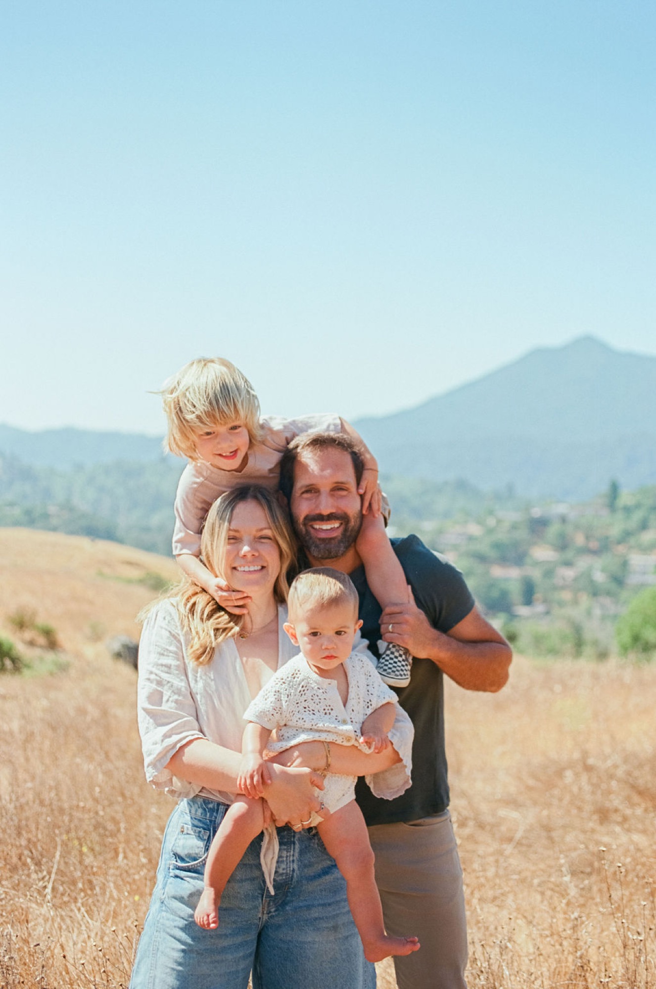 A bearded father grins broadly while holding his youngest, as his wife and two blonde children cluster close on sun-drenched golden hills beneath distant mountains