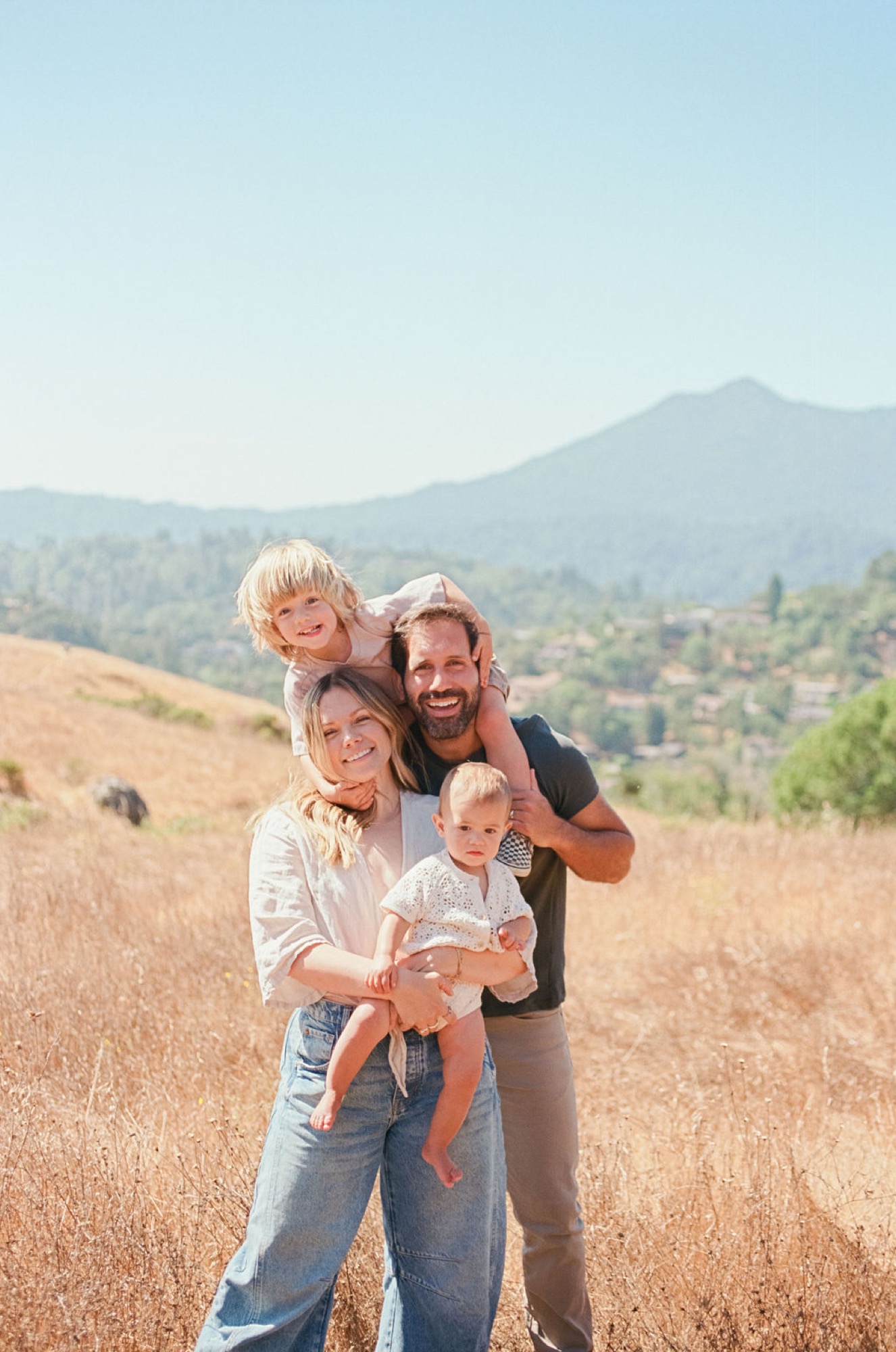 A bearded father grins while holding his toddler, his wife smiling beside him with their blonde child perched on his shoulders in golden hillside grass