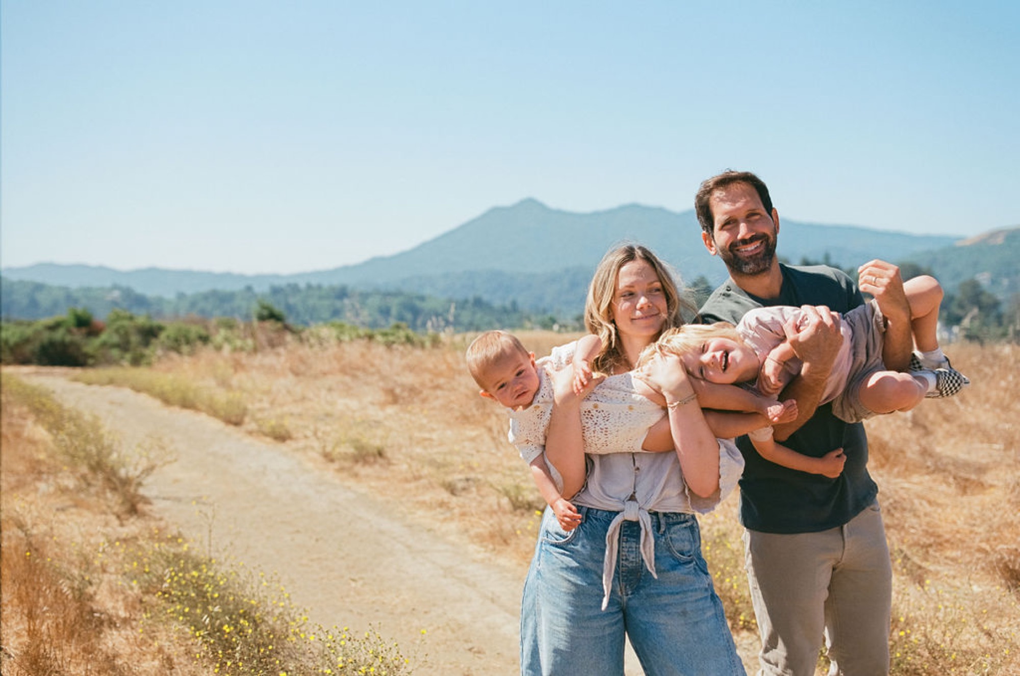 Parents playfully lift their two young children in a sun-drenched meadow, with rolling forested mountains rising behind them under clear blue sky