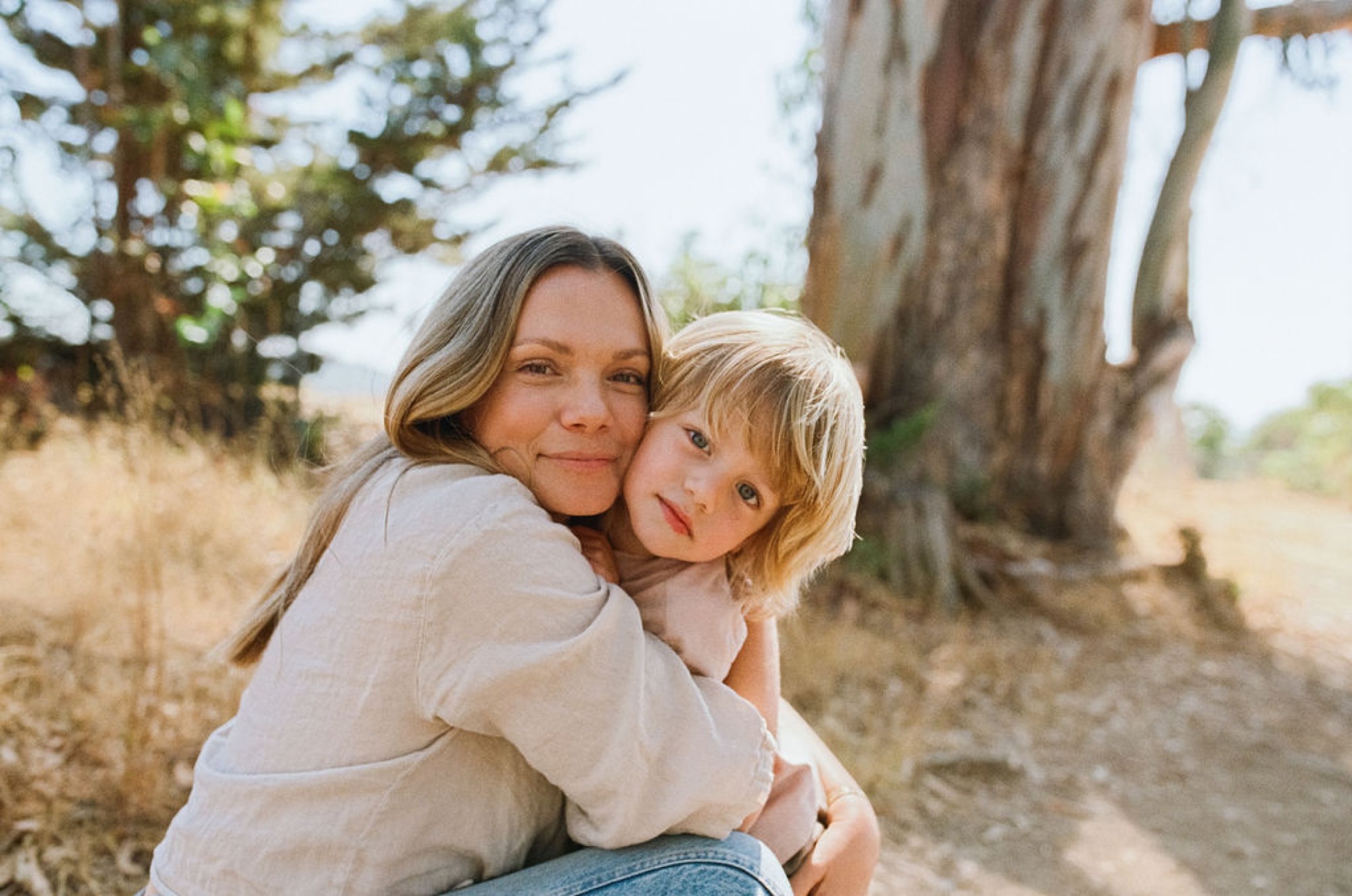 A mother carries her young blonde child on her back along a dusty trail, both gazing warmly toward the camera with golden afternoon light filtering through towering cypress trees