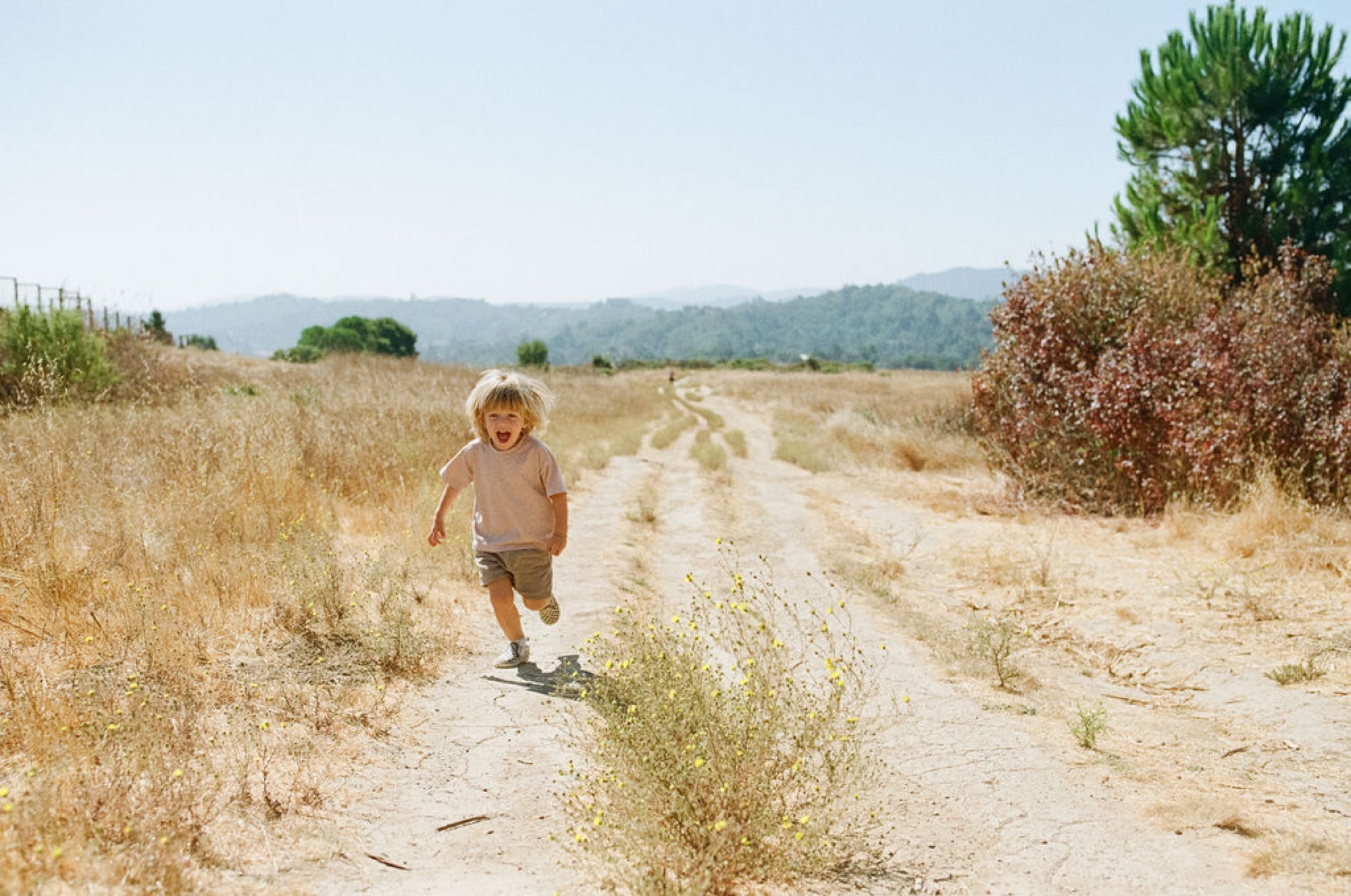 A blonde toddler runs joyfully down a dusty rural path, mouth open in delight, with dry golden grassland and tree-lined hills stretching behind