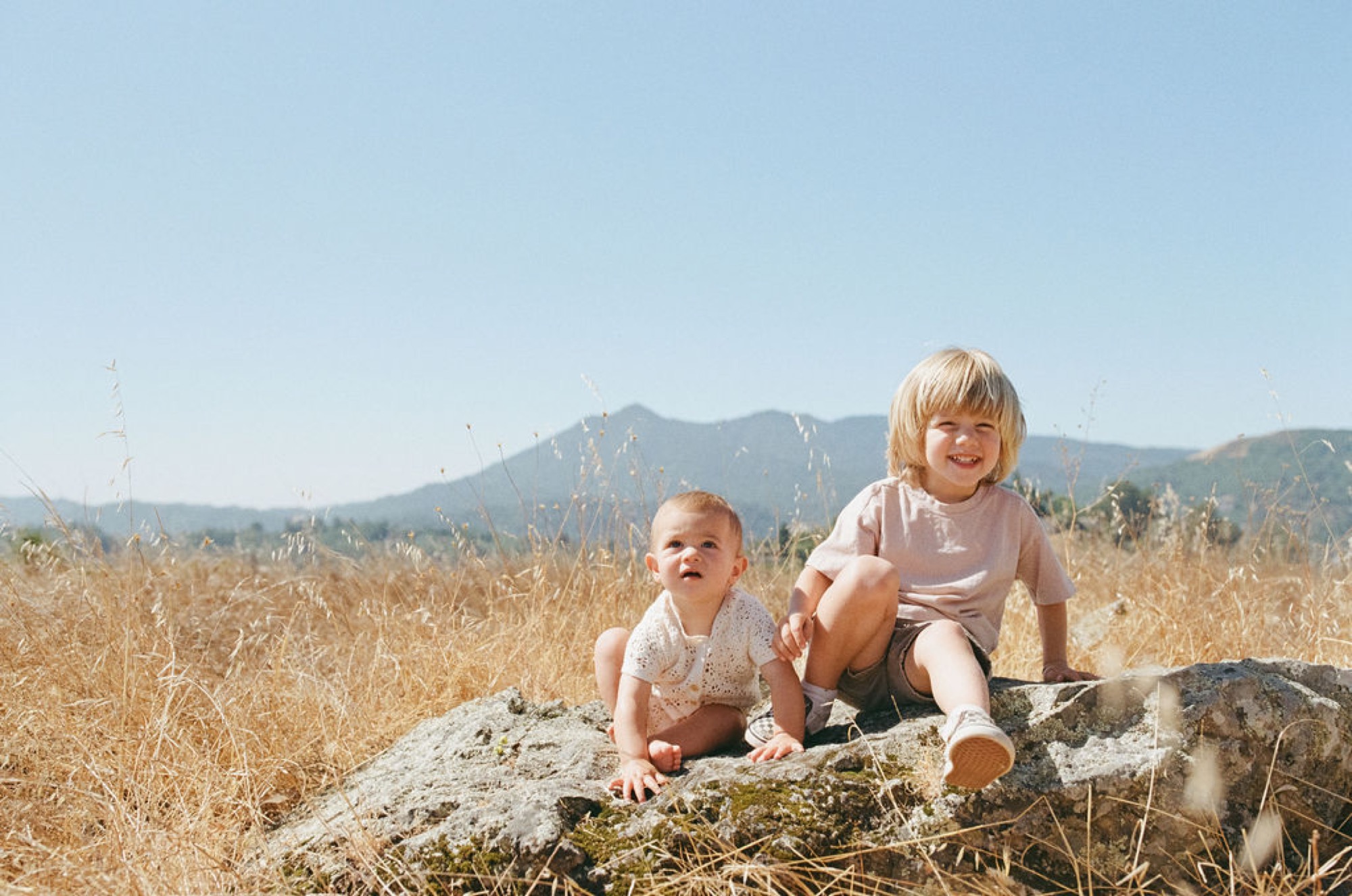 Two young children sit on a sun-warmed boulder in a golden grassland, the older sibling beaming while the infant gazes upward at distant mountains