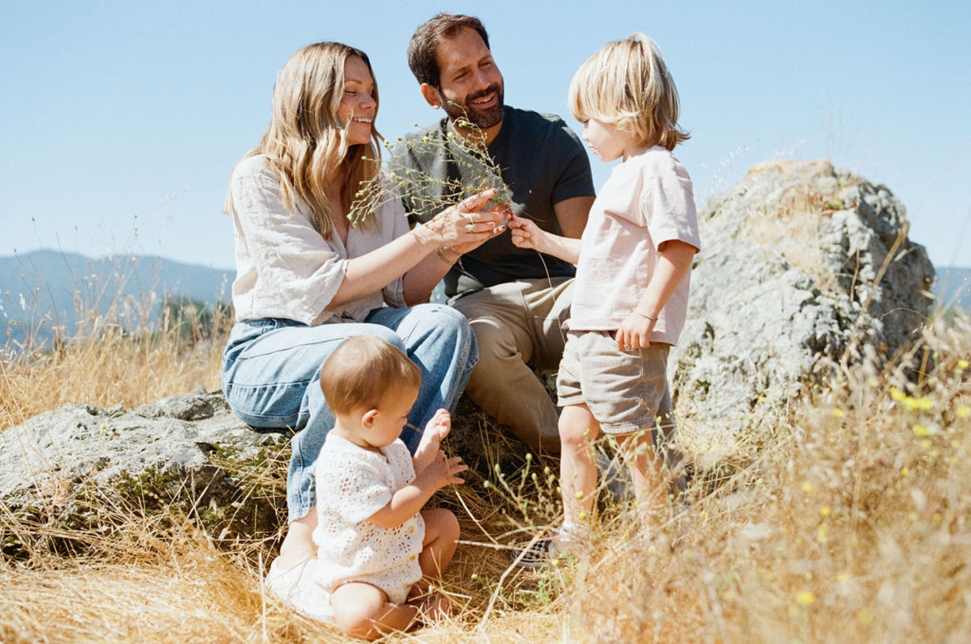 A family of four crouches together on a sun-lit hillside, examining dried grasses and plants with genuine curiosity and joy