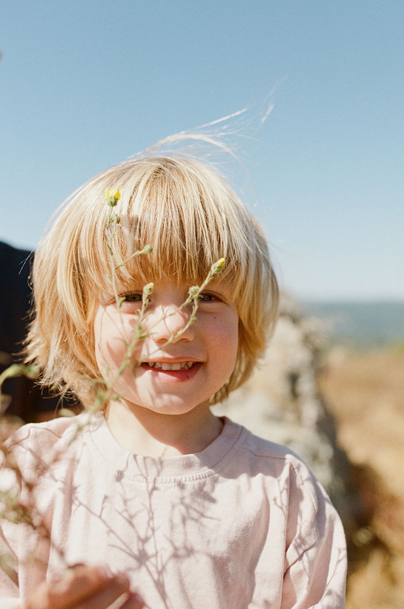 A blonde-haired child grins broadly at the camera while holding delicate green wildflower stems, squinting in bright sunlight against a blurred coastal hillside
