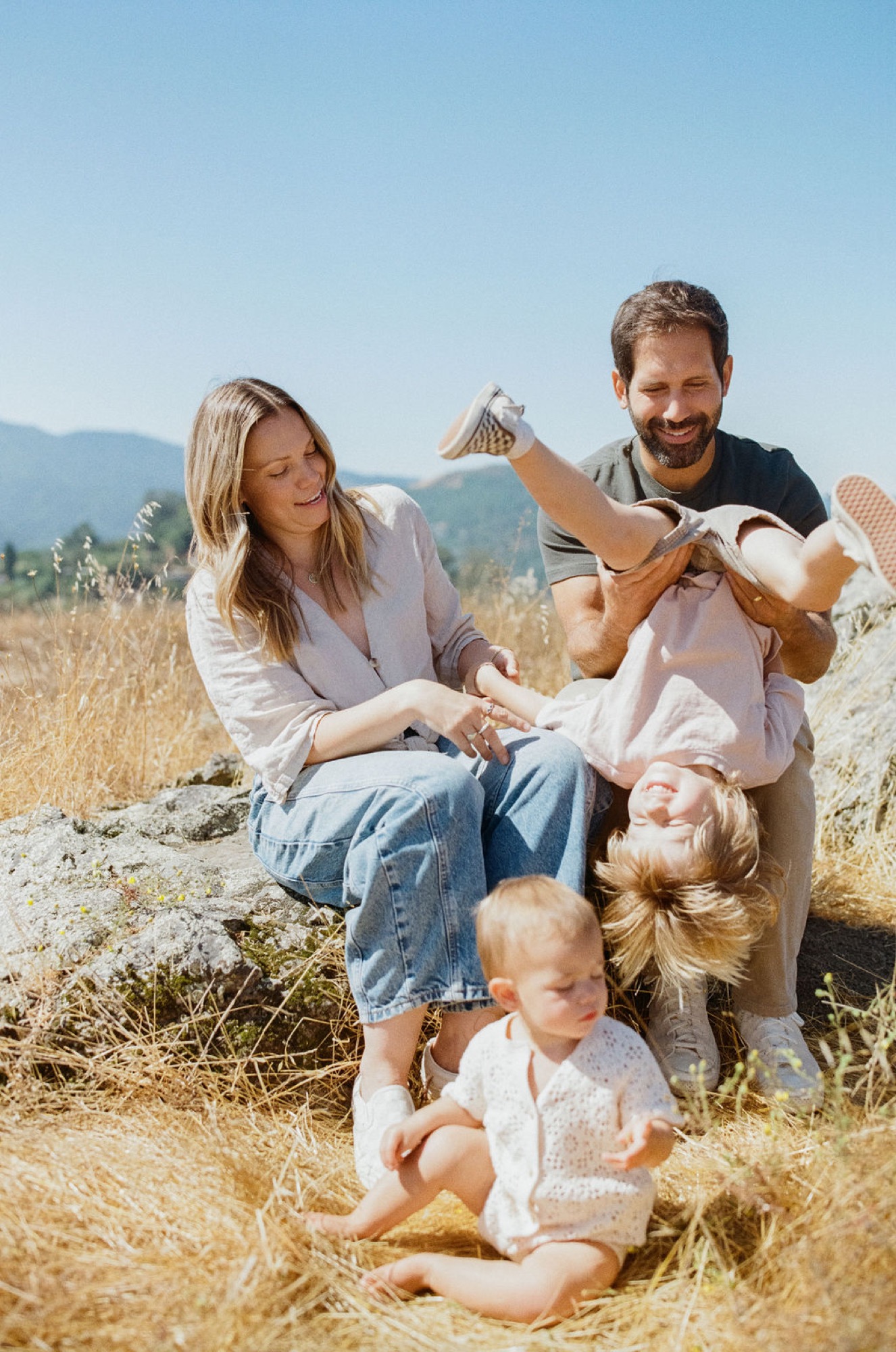 A family of four shares a joyful moment on sun-warmed earth, with two toddlers laughing while parents guide them beneath clear skies and distant hills
