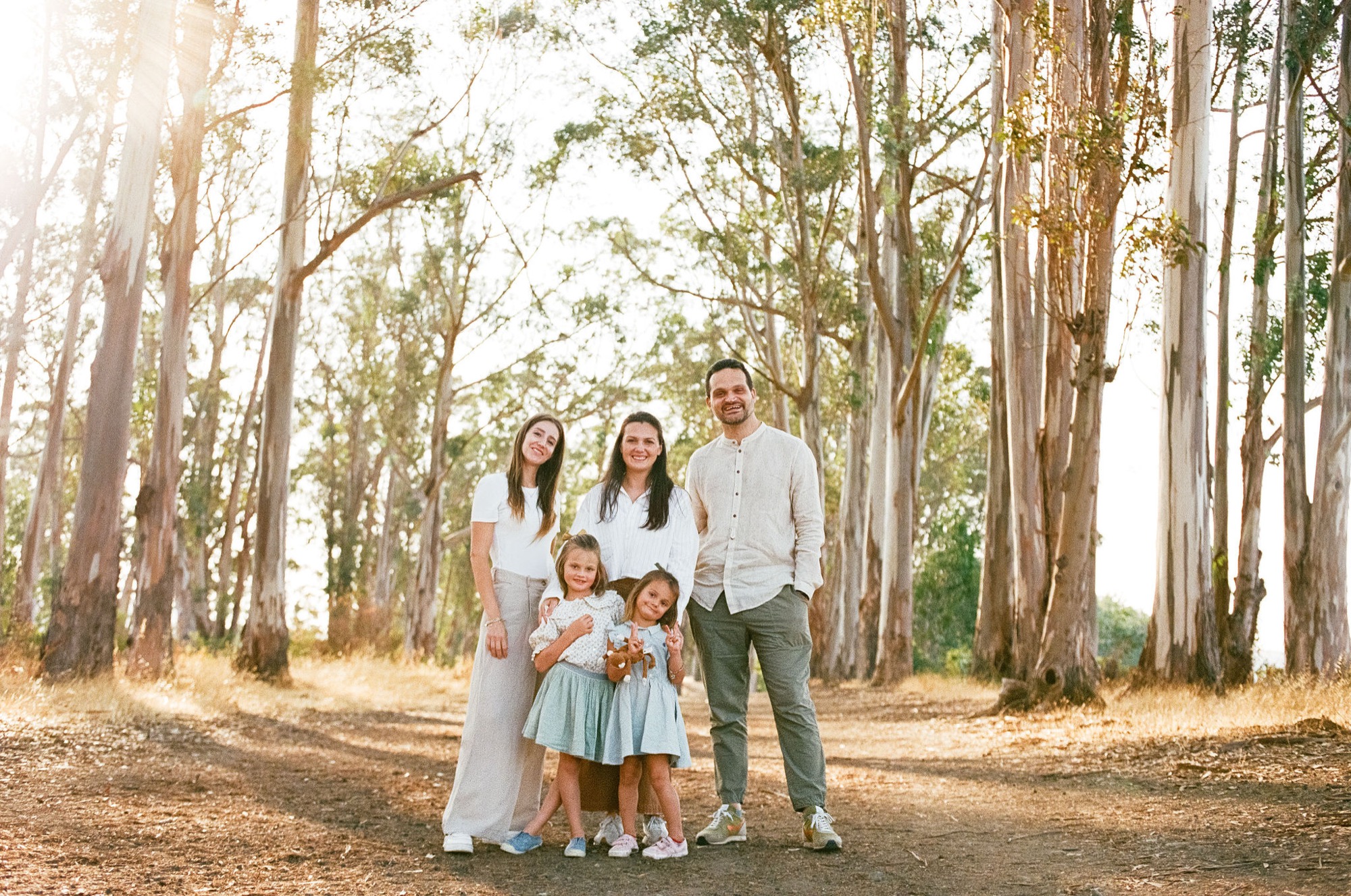 Family of five stands together on a shaded dirt path lined with tall eucalyptus trees, bathed in warm golden sunlight filtering through the canopy